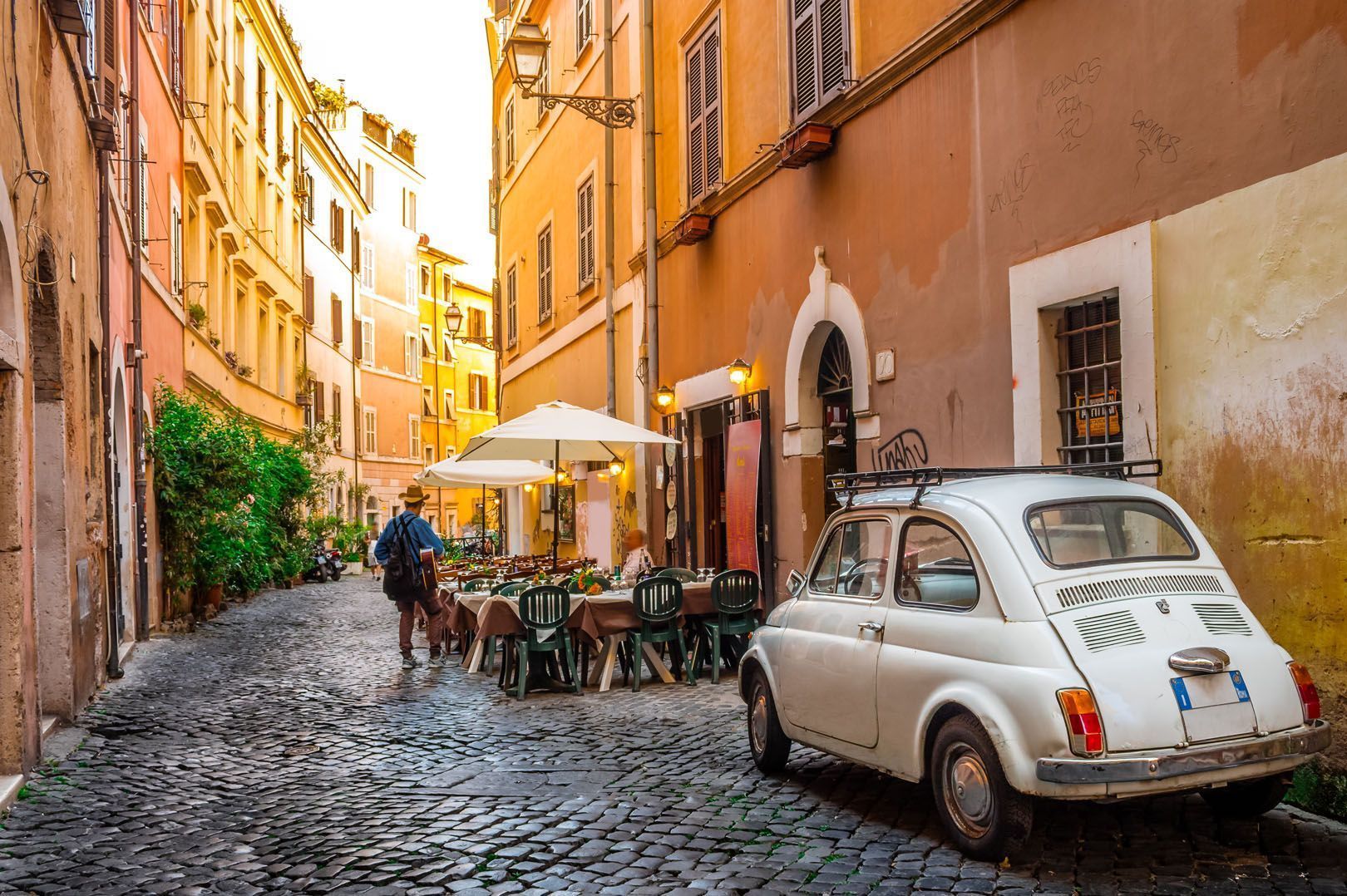 A vintage white car parked on a cobblestone street next to an outdoor cafe, with a man carrying a guitar walking by.