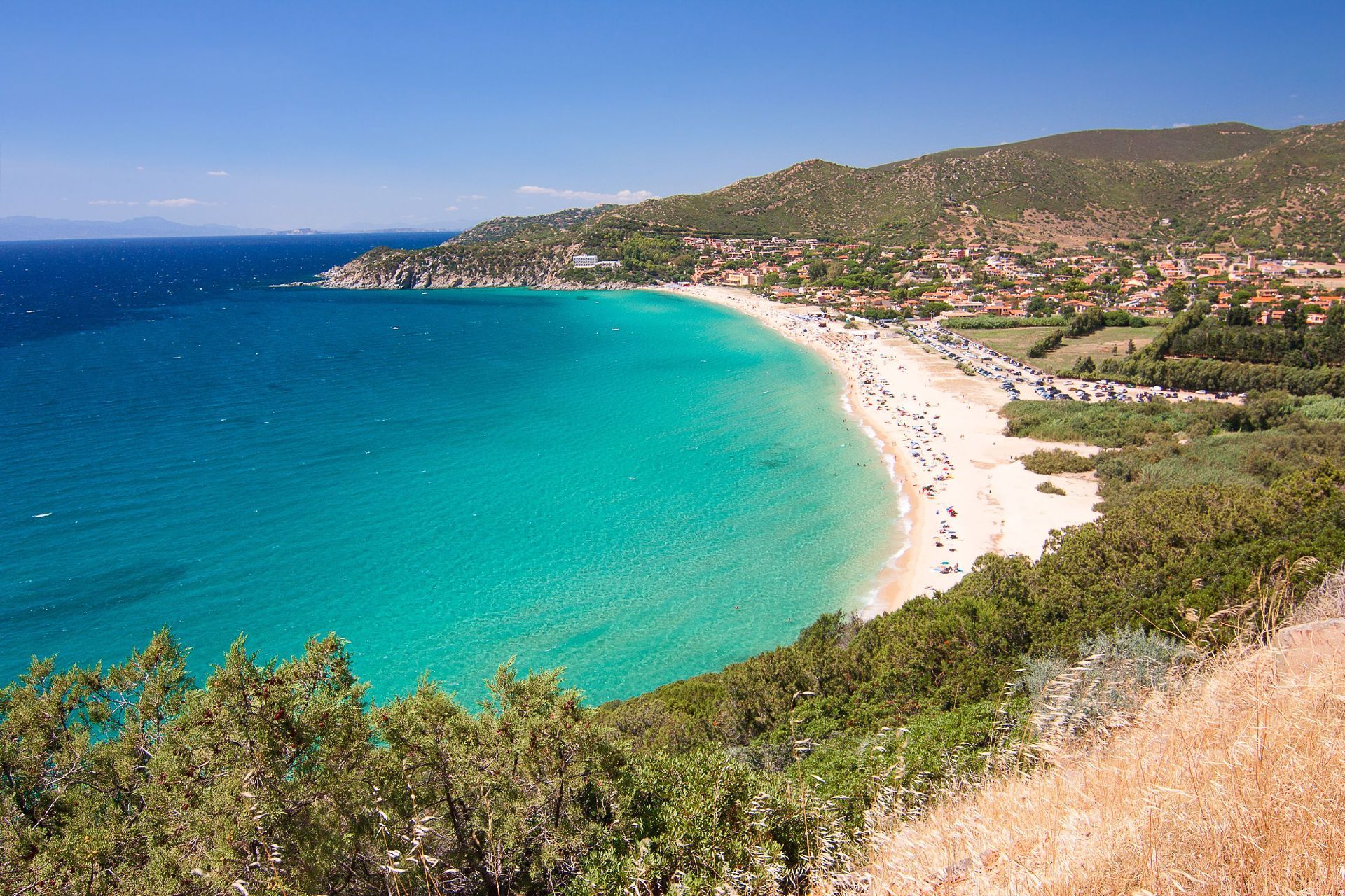 Una vista aerea di una spiaggia sabbiosa affollata e a forma di mezzaluna con acqua turchese, una città costiera e verdi colline sotto un cielo azzurro chiaro.