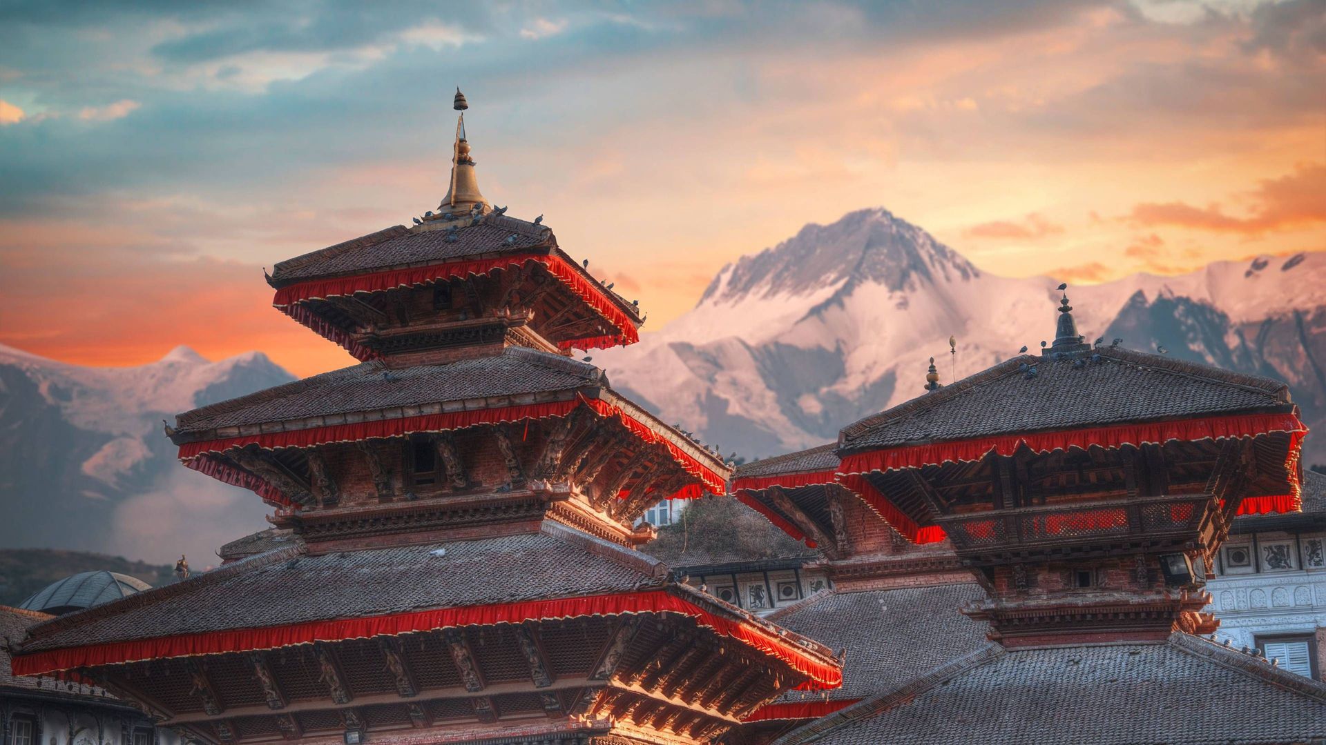 Mehrstufige Pagodendächer mit roter Zierleiste stehen vor schneebedeckten Bergen unter einem farbenfrohen Sonnenuntergangshimmel.