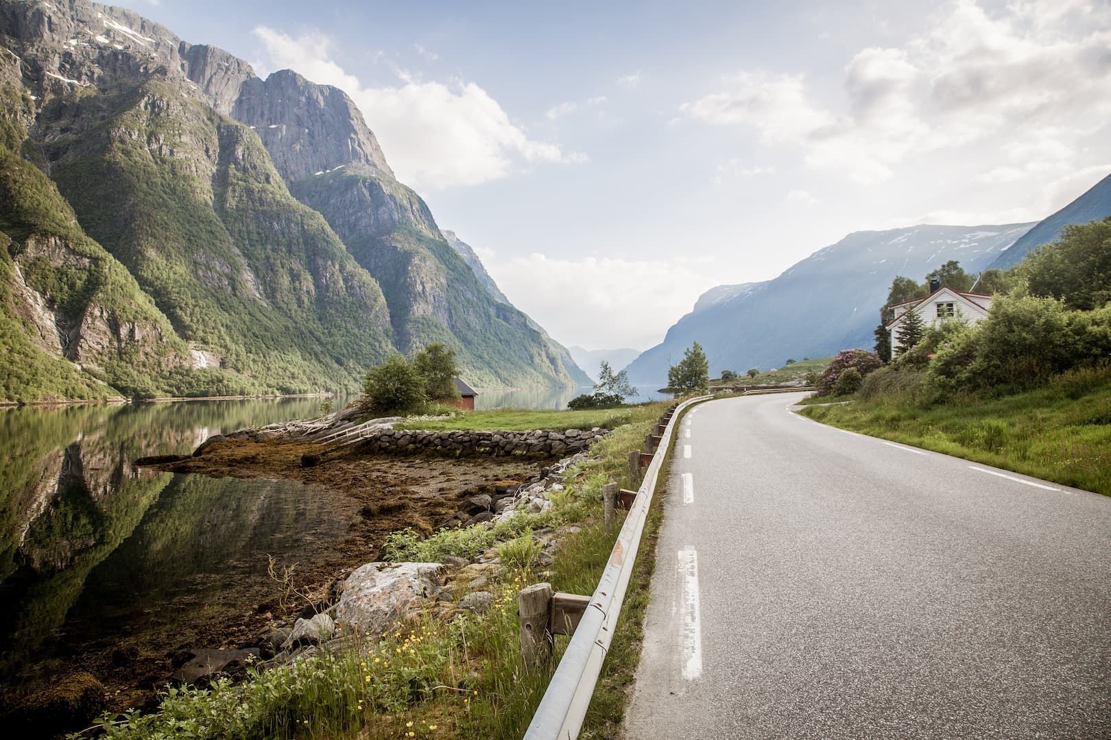 Une route goudronnée serpente le long d'un fjord, avec des montagnes vertes et abruptes s'élevant de l'eau sous un ciel partiellement nuageux.