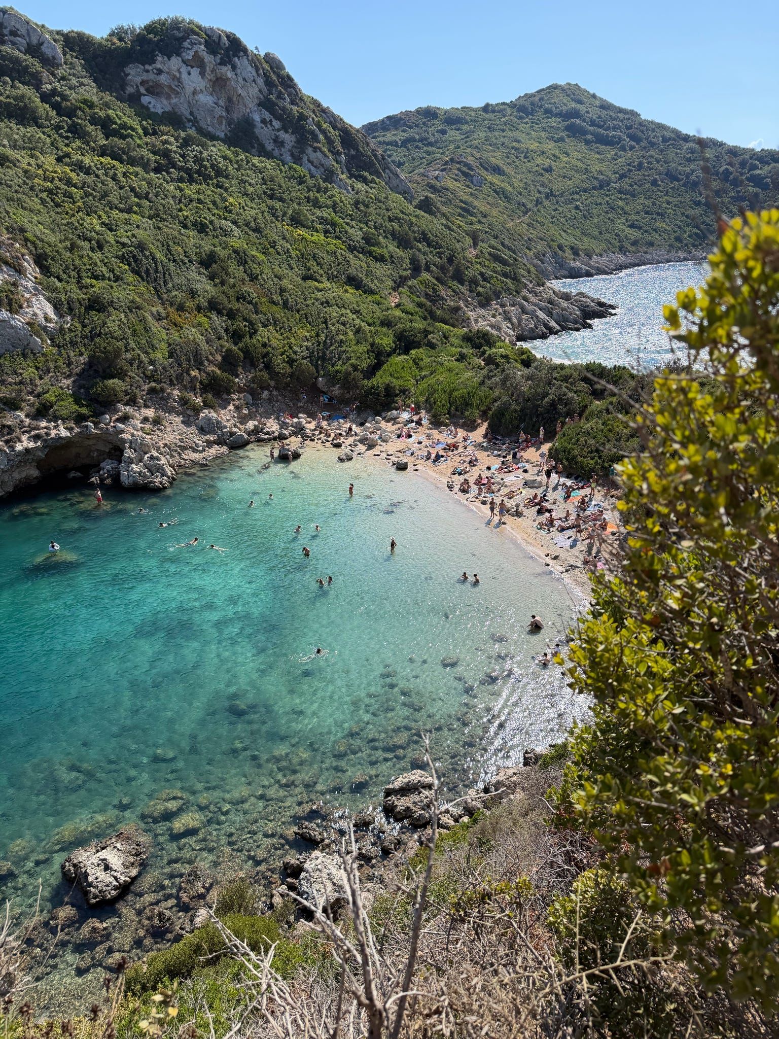 Des personnes nagent et bronzent dans une crique isolée aux eaux turquoise entourée de collines verdoyantes.