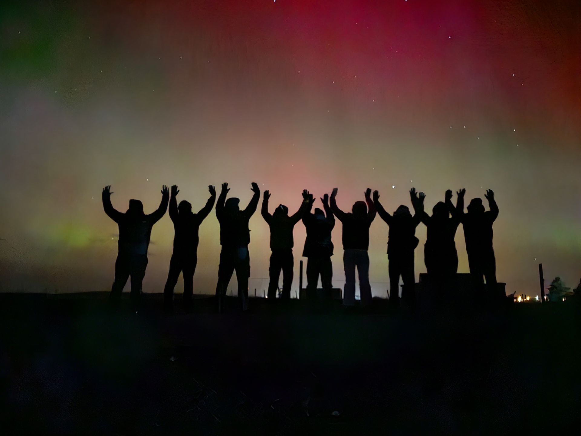 A WeRoad group trip standing with raised arms in silhouette under colorful aurora lights in the night sky.