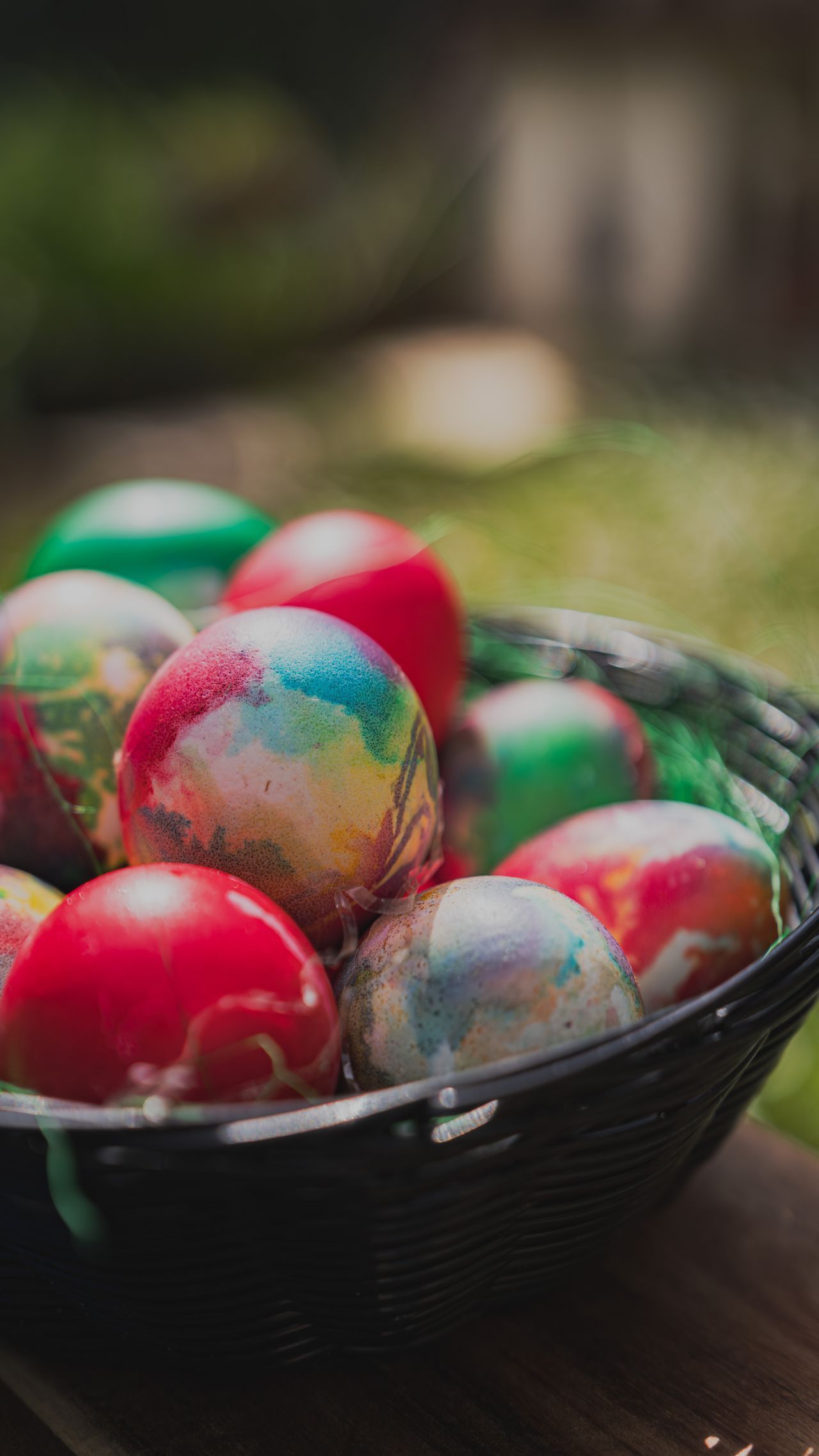 Uova di Pasqua dipinte colorate raccolte dentro un cesto di vimini scuro all'aperto su una superficie di legno.