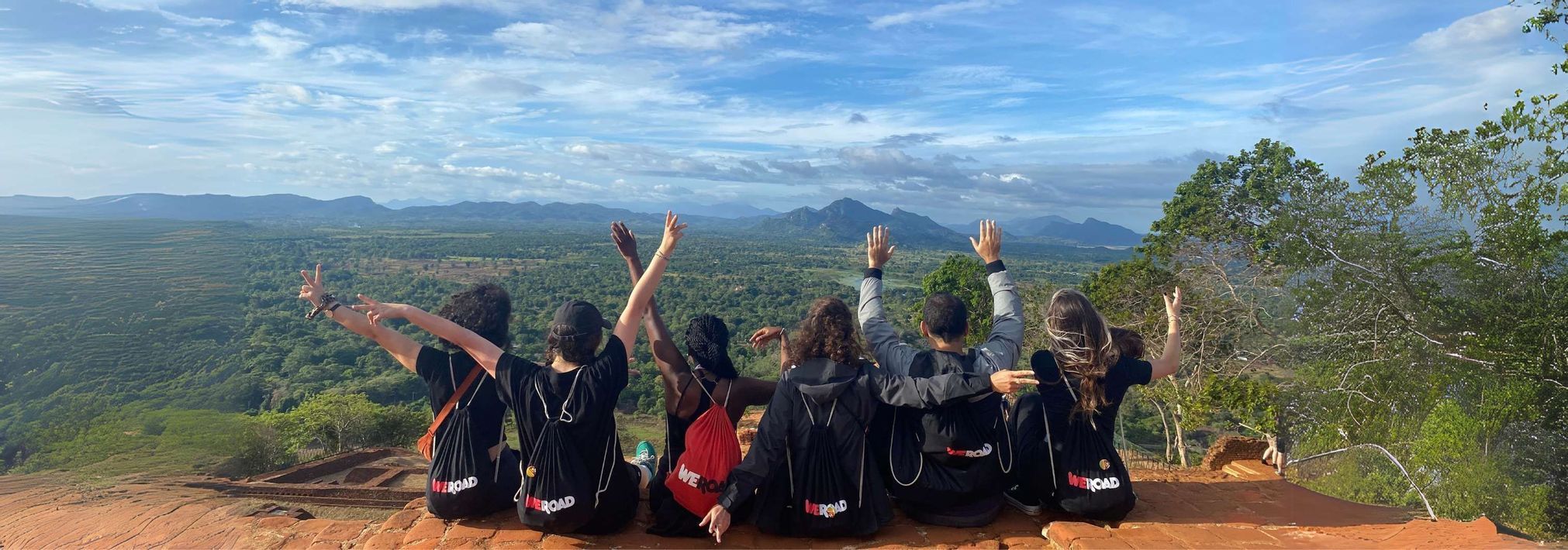 Un gruppo di viaggio WeRoad siede insieme con le braccia alzate davanti a un'ampia valle verde e montagne lontane.