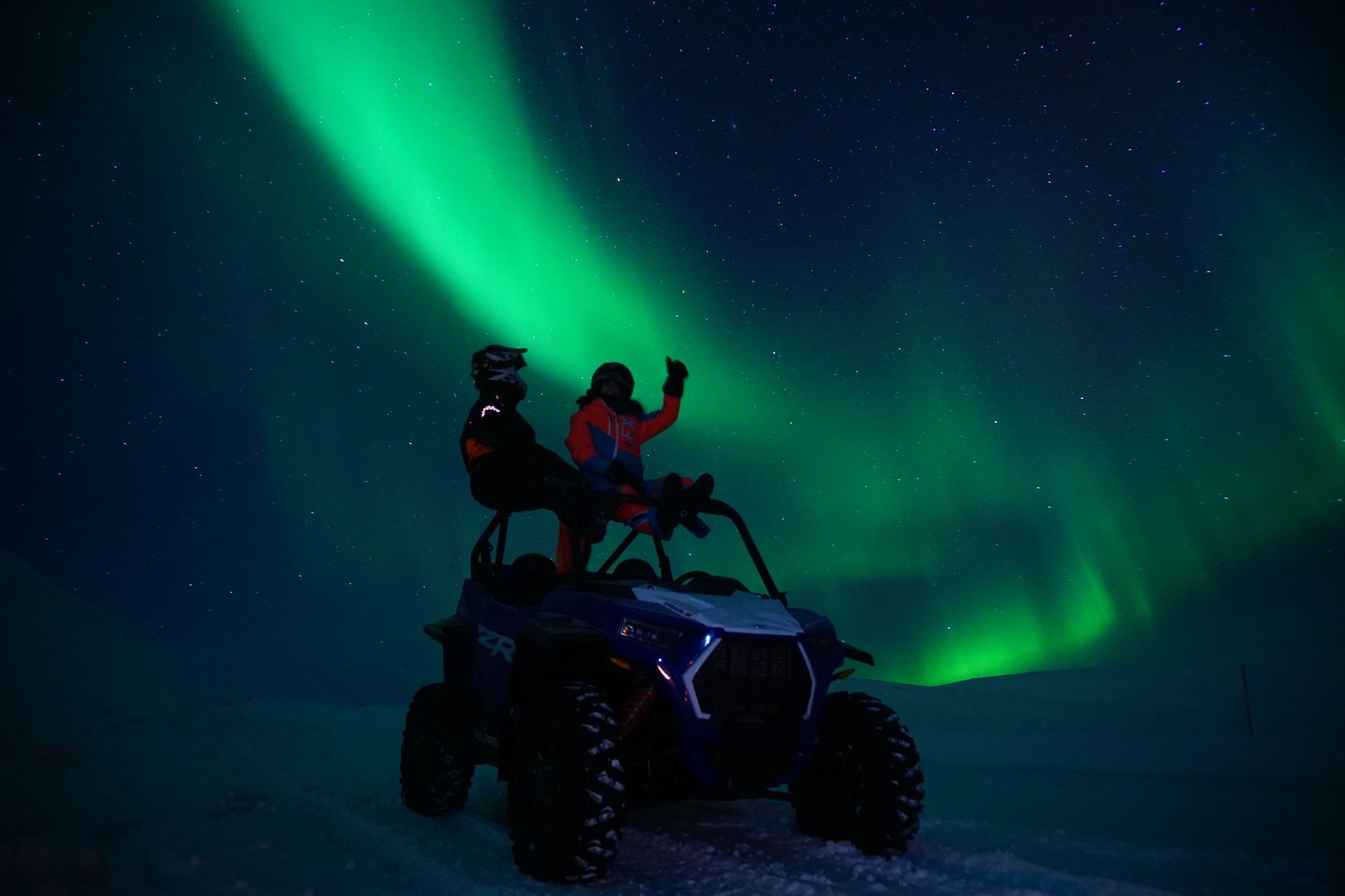 Due persone con abbigliamento invernale sedute sopra un buggy fuoristrada sotto una brillante aurora boreale verde in un cielo stellato notturno.