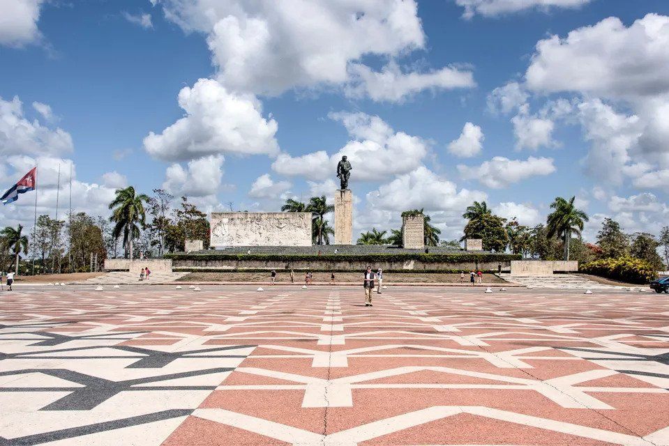 A wide-angle view of a monument with a statue overlooking a large plaza with a red, white, and black geometric pattern under a cloudy blue sky.