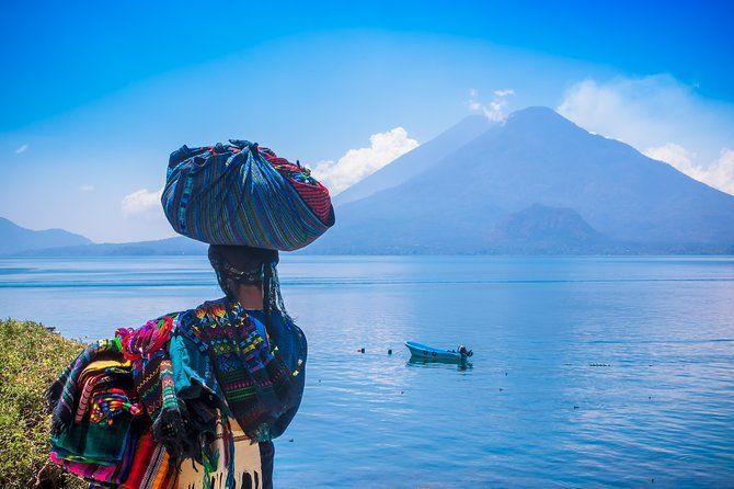 A person in colorful traditional clothing carries a bundle on their head, standing by a lake with large mountains in the background.