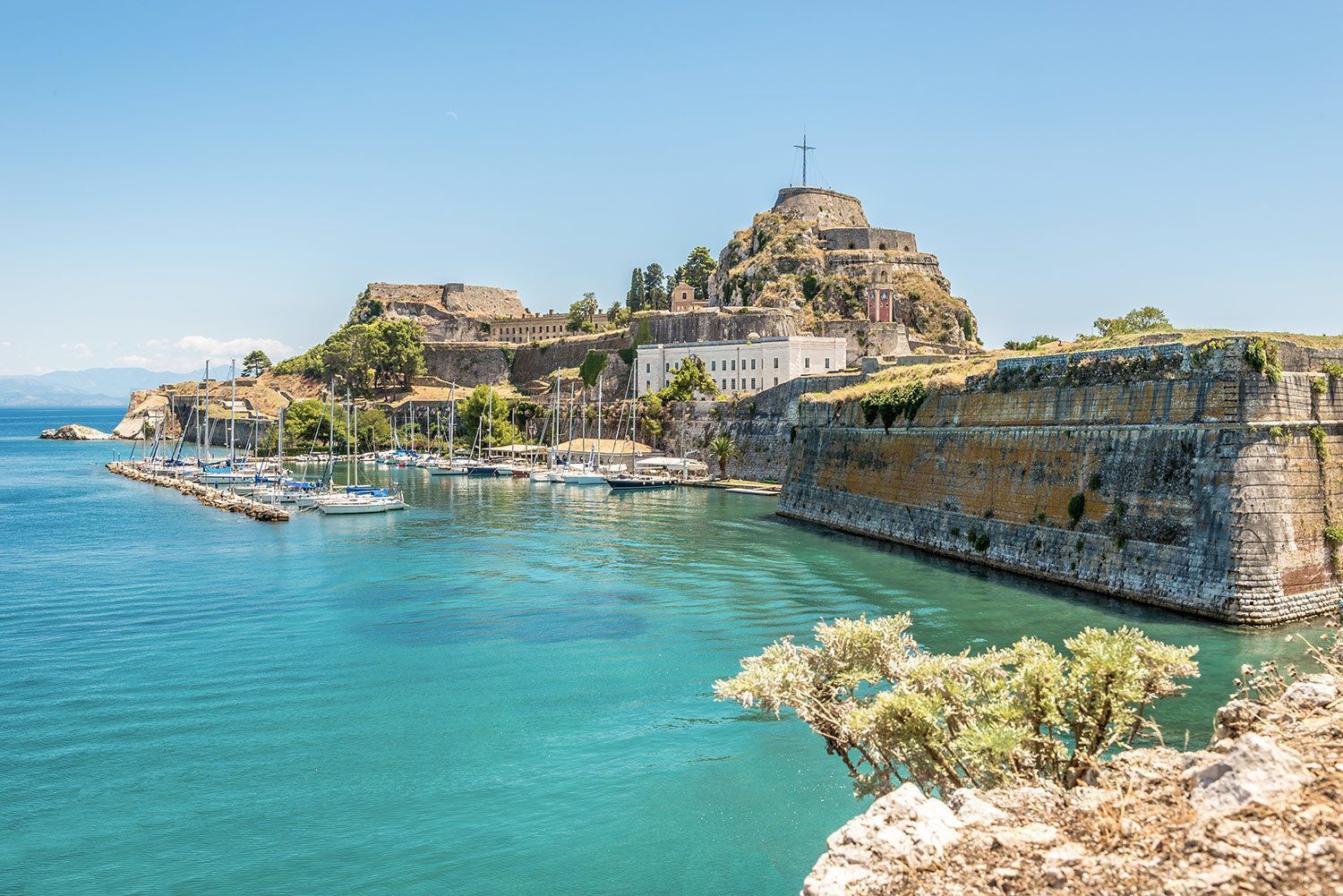 A stone fortress on a hill overlooks a marina with sailboats docked in a bay with turquoise water on a sunny day.