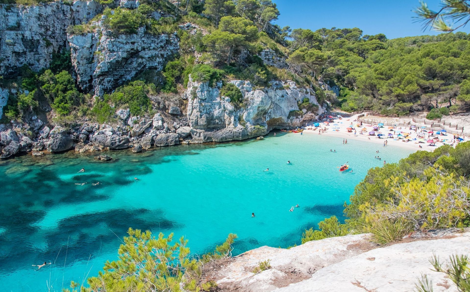 Vue aérienne d'une crique où des personnes nagent dans une eau turquoise claire et se détendent sur une plage de sable blanc entourée de falaises rocheuses et d'arbres.