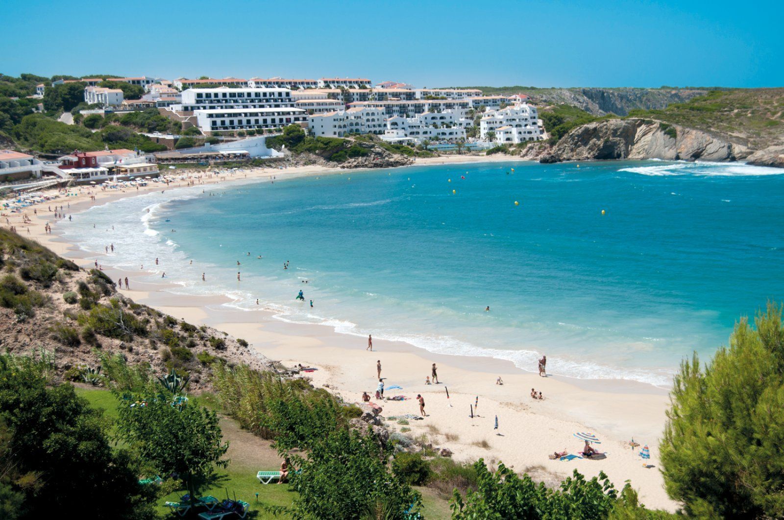 Une large vue d'une crique de sable où l'on se baigne dans des eaux turquoise, avec des bâtiments blancs sur les falaises au-dessus de la plage.