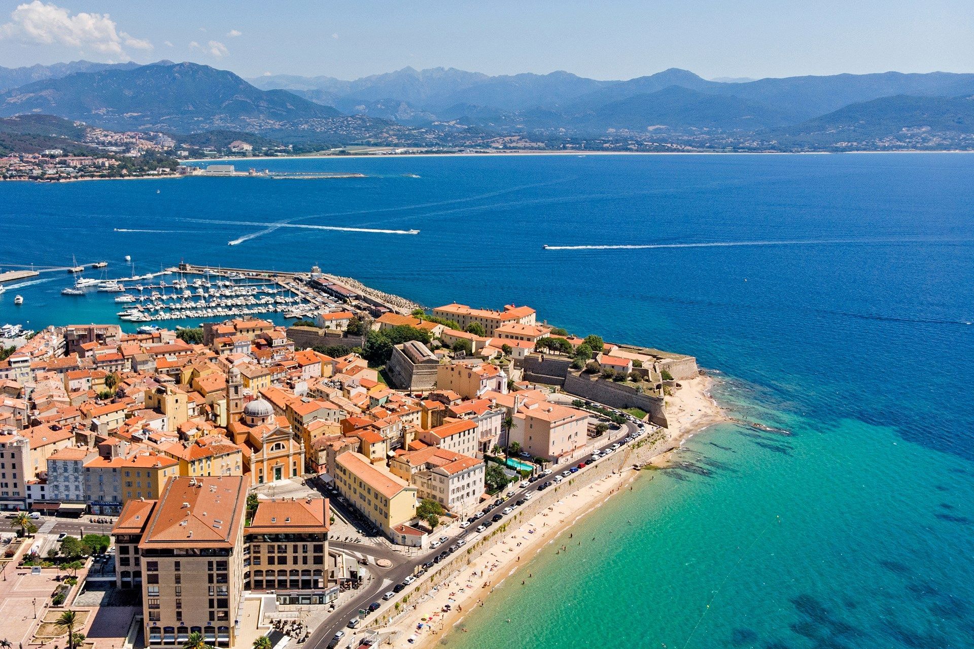 Vue aérienne d'une ville côtière avec un port, une plage de sable et une mer turquoise, avec des montagnes lointaines sous un ciel bleu.