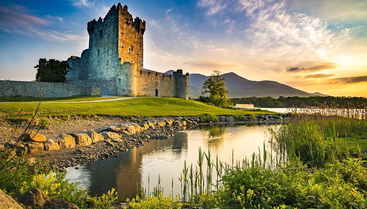 Un castillo de piedra en la orilla herbosa de un lago tranquilo, iluminado por el sol poniente, con montañas a lo lejos.
