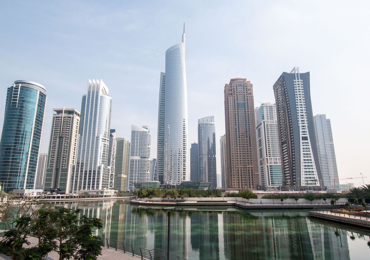 A modern city skyline with tall glass skyscrapers reflecting in a calm body of water in the foreground.