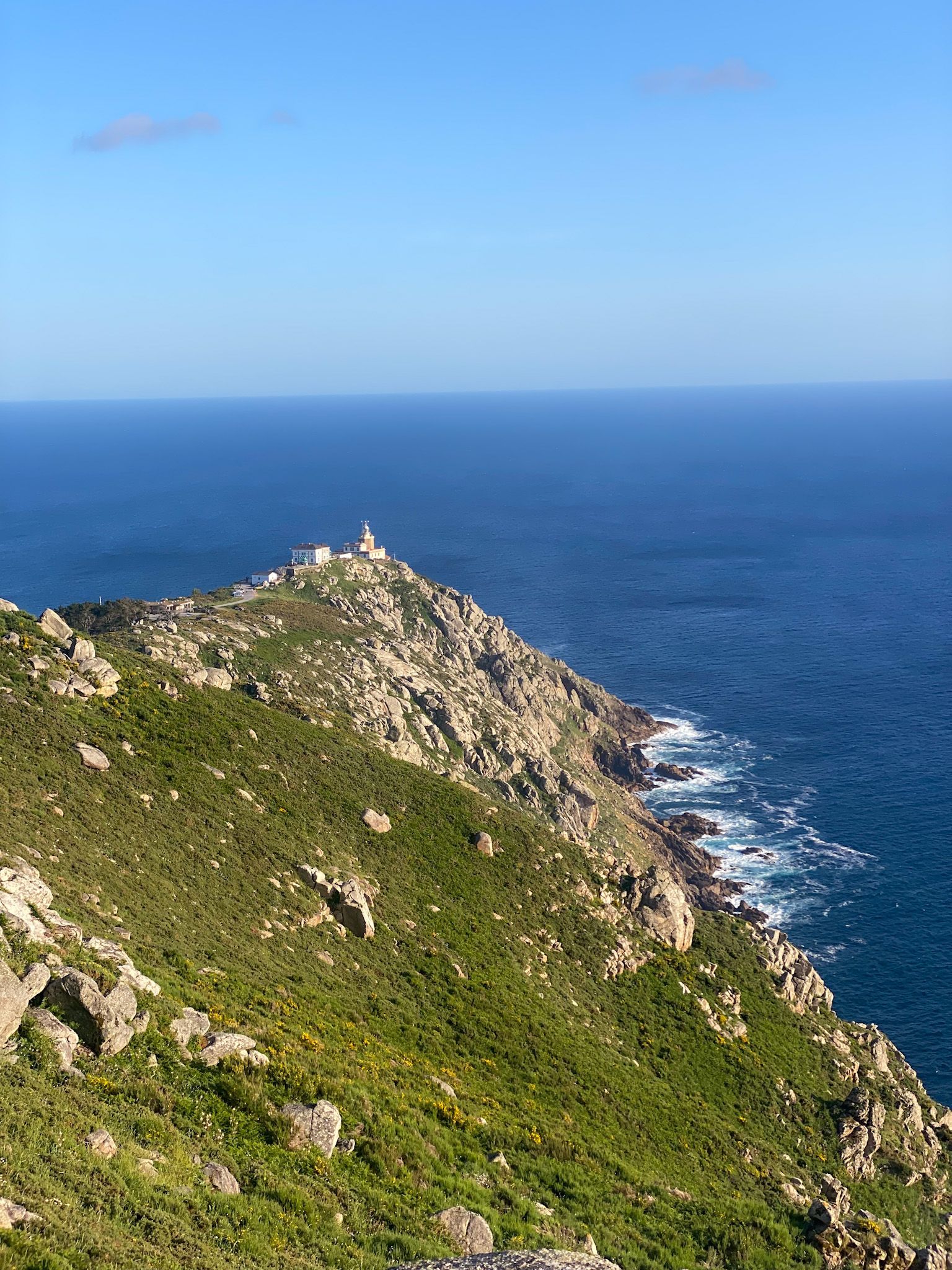 Un faro y edificios adyacentes se asientan en la punta de una península rocosa y verde, con vistas a un vasto océano azul bajo un cielo despejado.