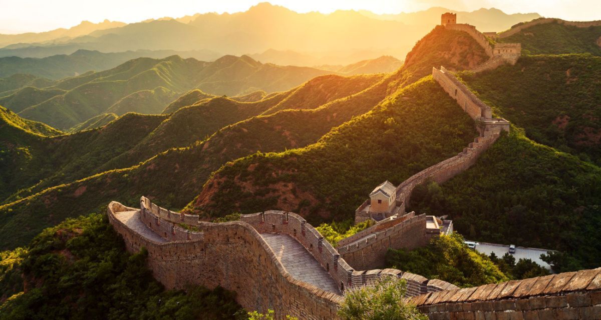 Eine lange Steinmauer mit Wachtürmen schlängelt sich über üppig grüne Berge im goldenen Licht eines Sonnenaufgangs.