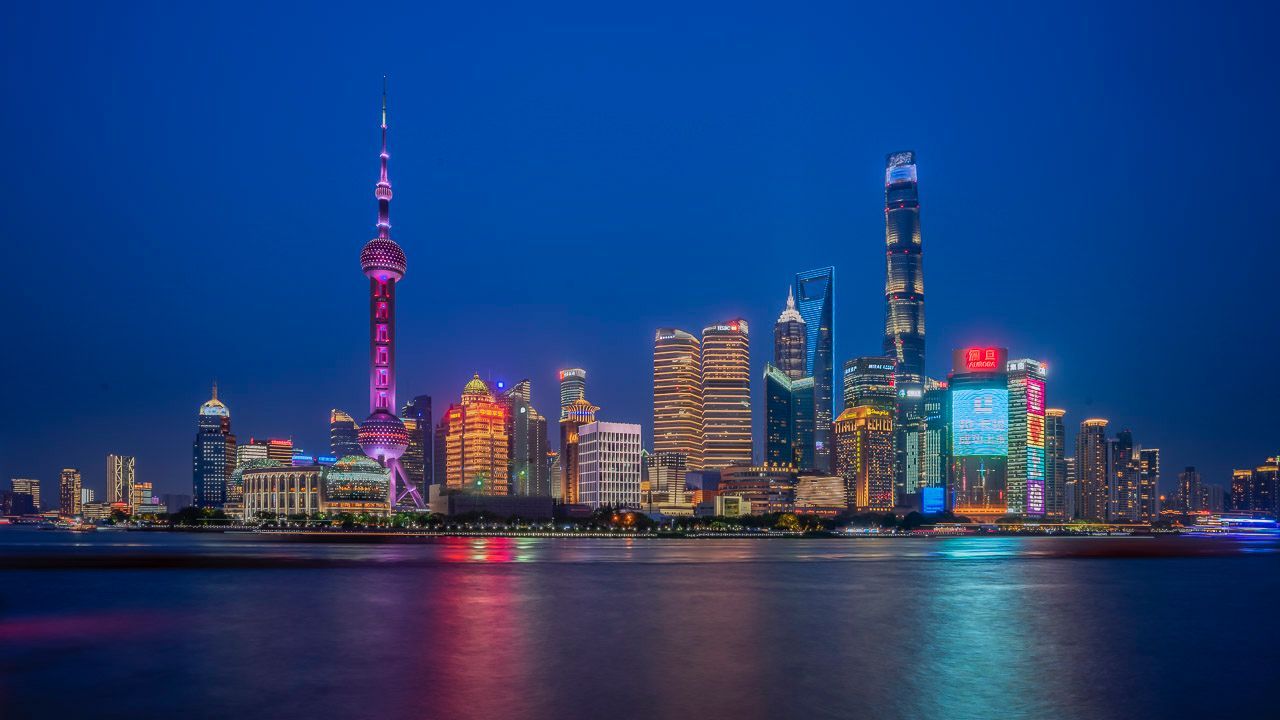 A modern city skyline at night, with illuminated buildings and a tall tower reflecting on the calm water.