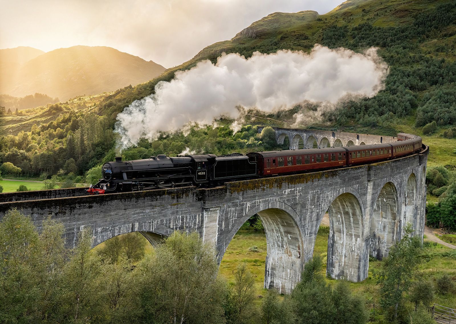 Un treno a vapore nero con carrozze rosse attraversa un viadotto in pietra curvilineo, emettendo fumo bianco in una valle verde e soleggiata.