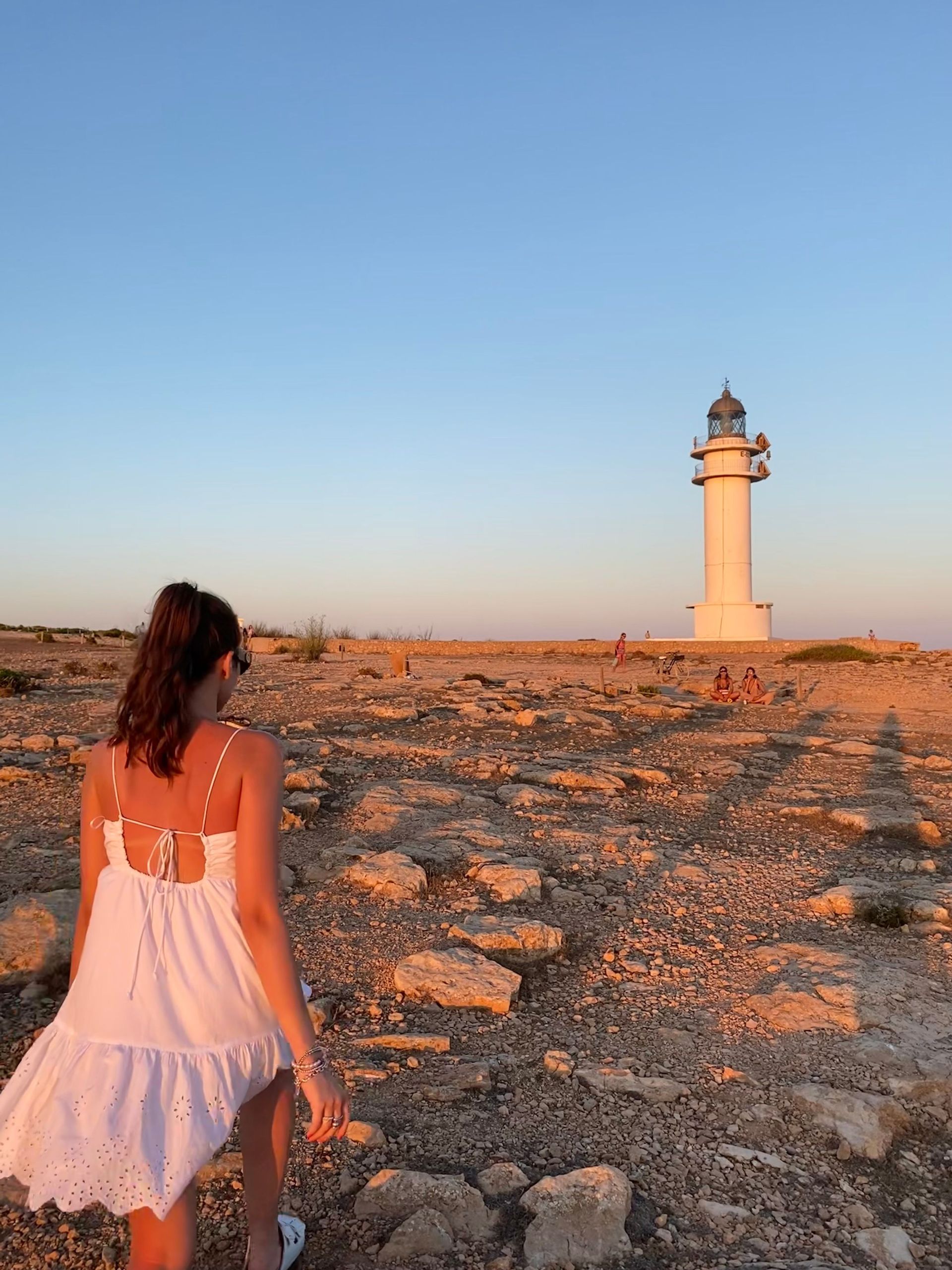 Una donna in abito bianco cammina attraverso un paesaggio roccioso verso un alto faro bianco durante un tramonto dorato.