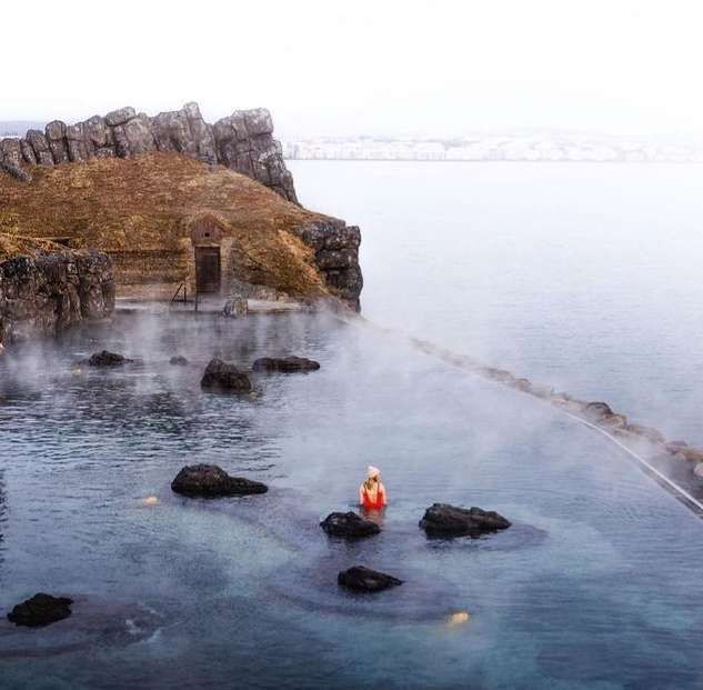 Eine Person in rotem Badeanzug und Beanie entspannt sich in einem dampfenden Infinity-Pool mit Blick auf ein größeres Gewässer neben einer felsigen Klippe.