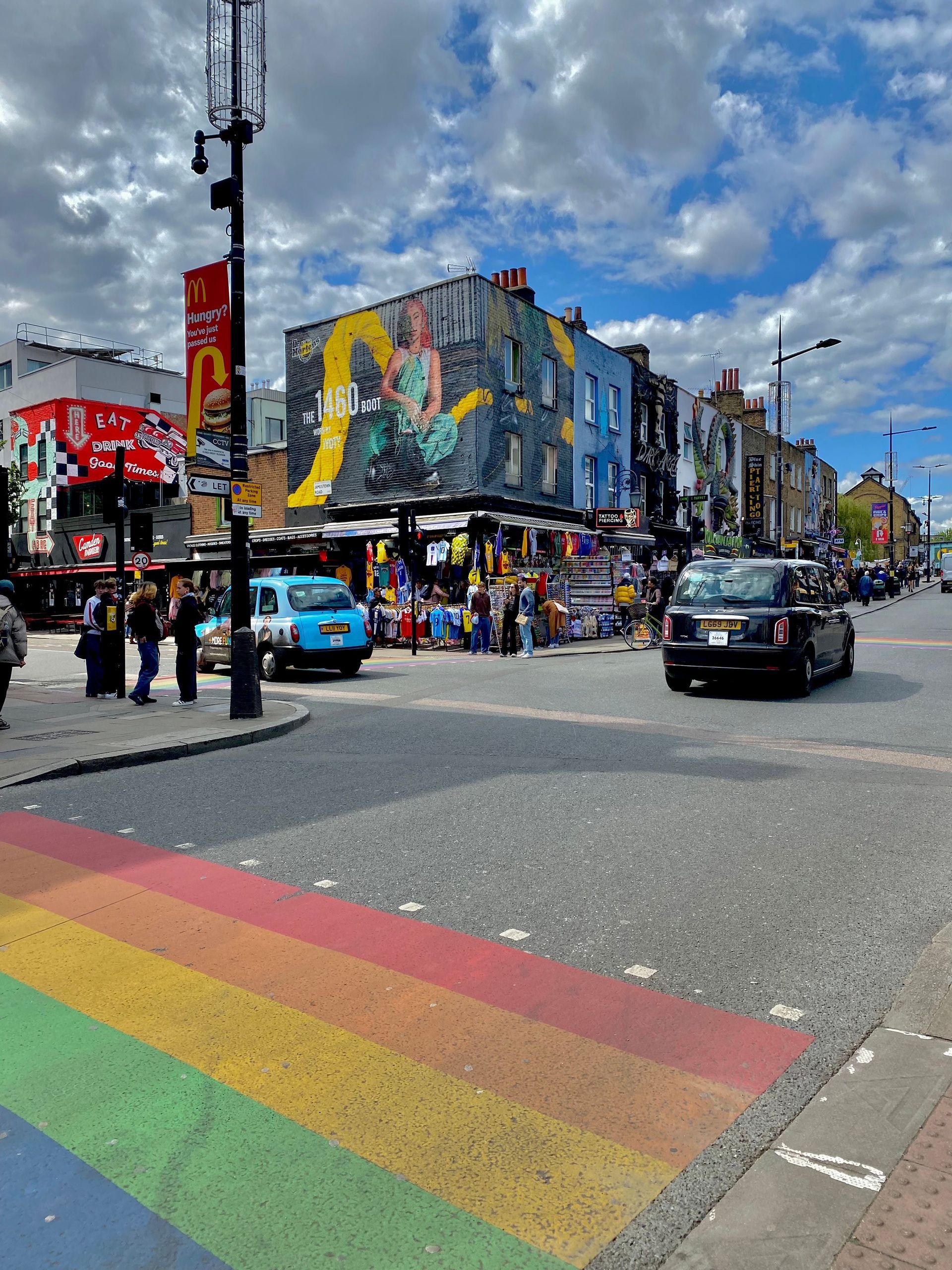 Un coin de rue en ville avec un passage piéton arc-en-ciel, des voitures, des piétons et des bâtiments recouverts de peintures murales colorées sous un ciel partiellement nuageux.