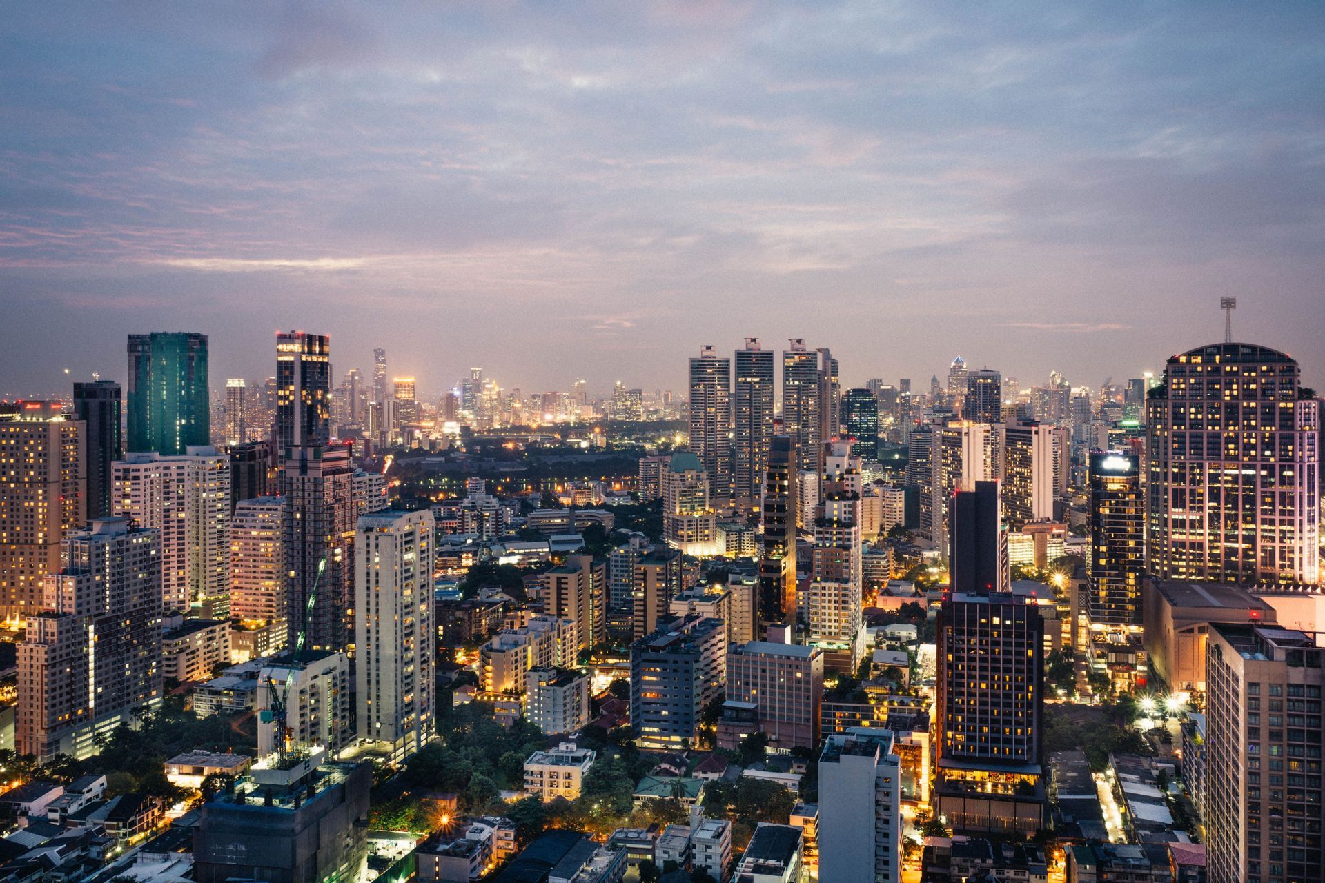 A sprawling city skyline with illuminated skyscrapers under a cloudy sky during twilight.