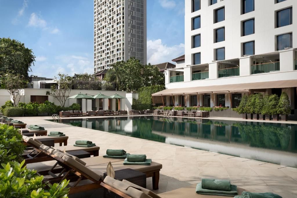 Empty lounge chairs with rolled green towels are lined up next to a long swimming pool with modern buildings in the background.