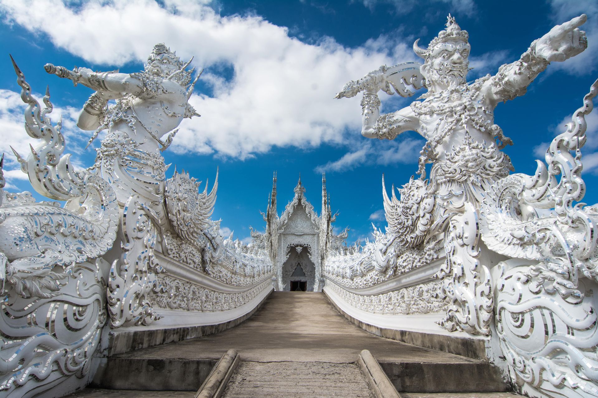A low-angle view of a walkway leading towards an ornate white temple, flanked by large, intricate statues under a blue sky.