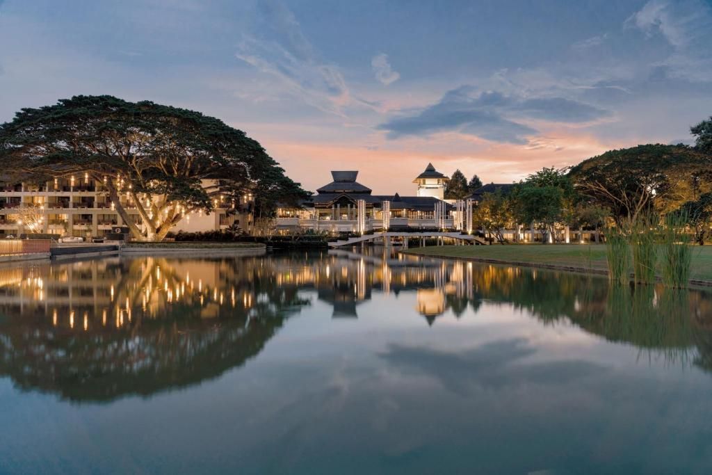An illuminated resort with traditional architecture reflects in a calm lake at dusk, alongside a large, decorated tree.