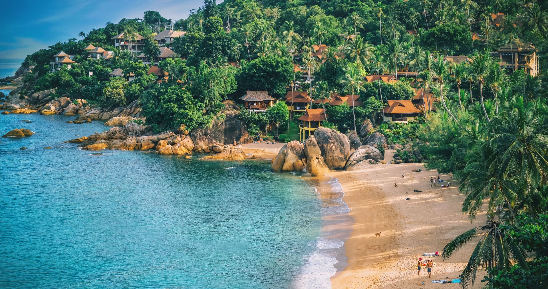 High-angle view of a sandy beach cove with turquoise water, large boulders, and villas nestled in a lush, green, palm-filled hillside.