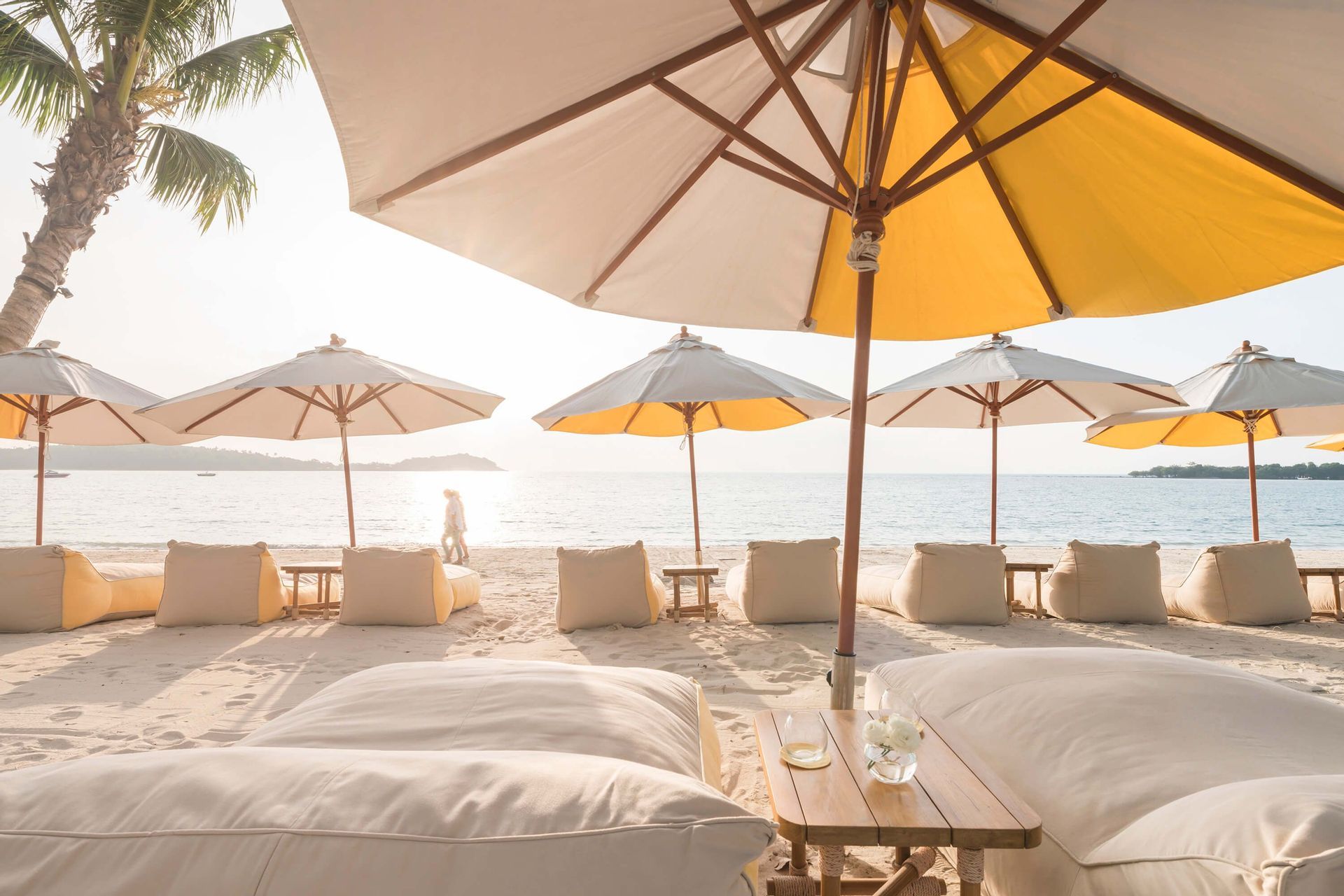 From under a large yellow and white umbrella, beanbag loungers are set up on a sandy beach facing the sea at sunrise.