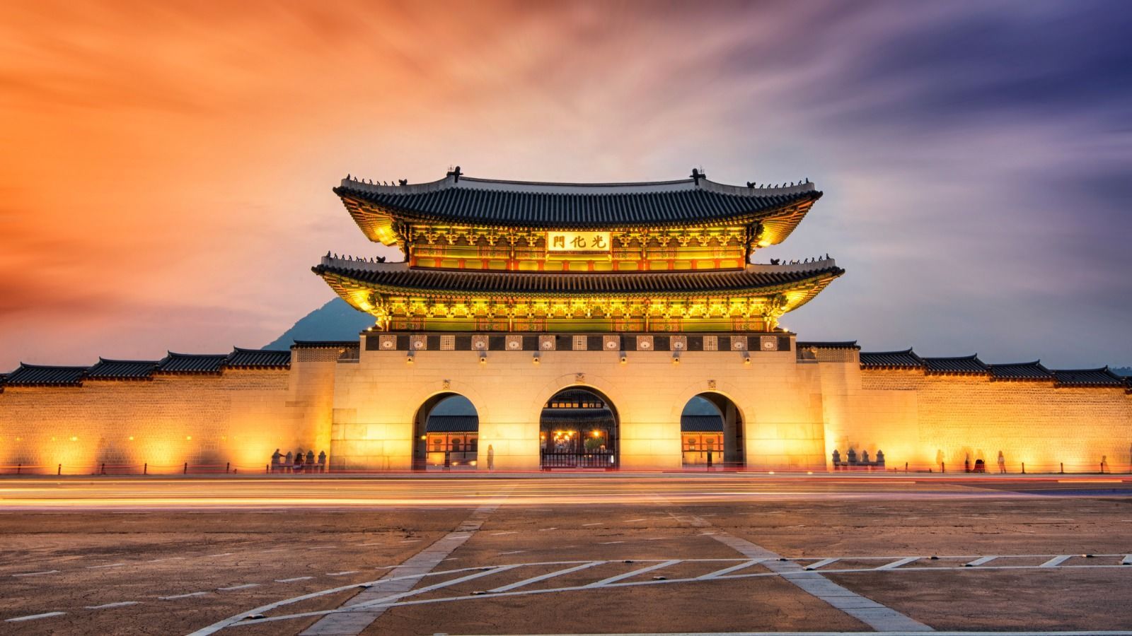 A traditional Asian gatehouse and its stone wall are brightly illuminated at dusk, with long streaks of traffic lights in the foreground.