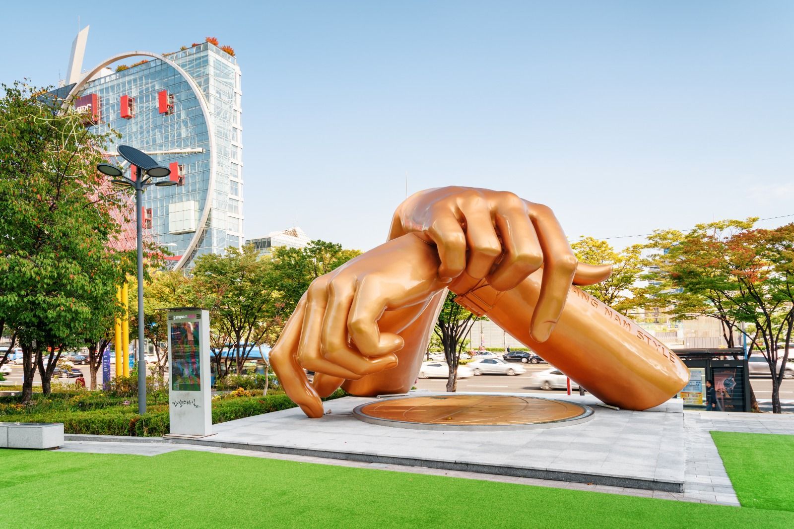 A large golden sculpture of two hands crossed in the 'Gangnam Style' dance pose sits in a city park in front of a modern skyscraper.