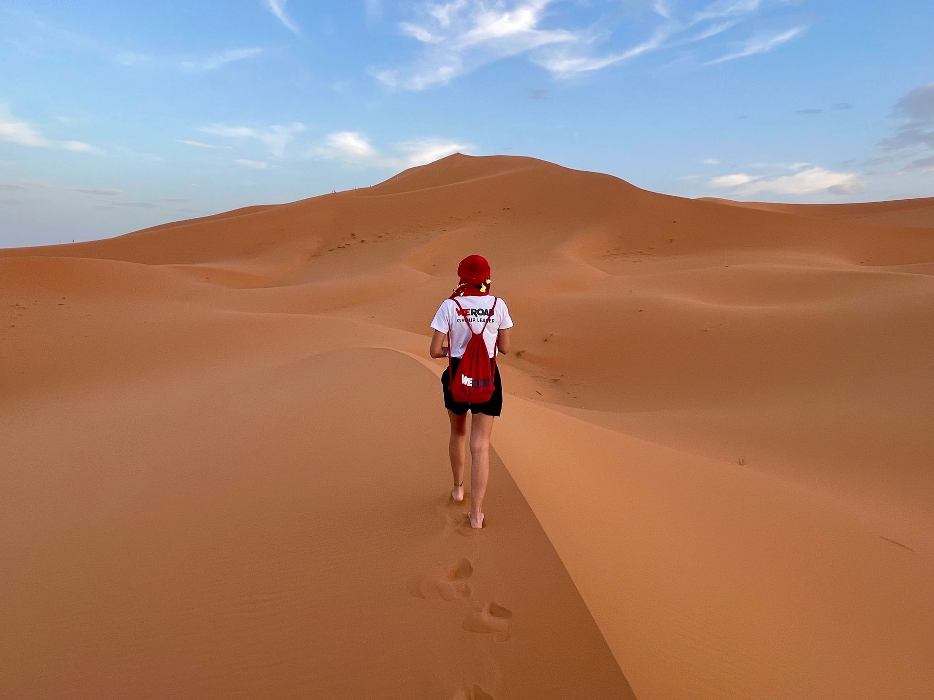 A person seen from behind wearing a WeRoad t-shirt and red cap walks barefoot up a large orange sand dune.