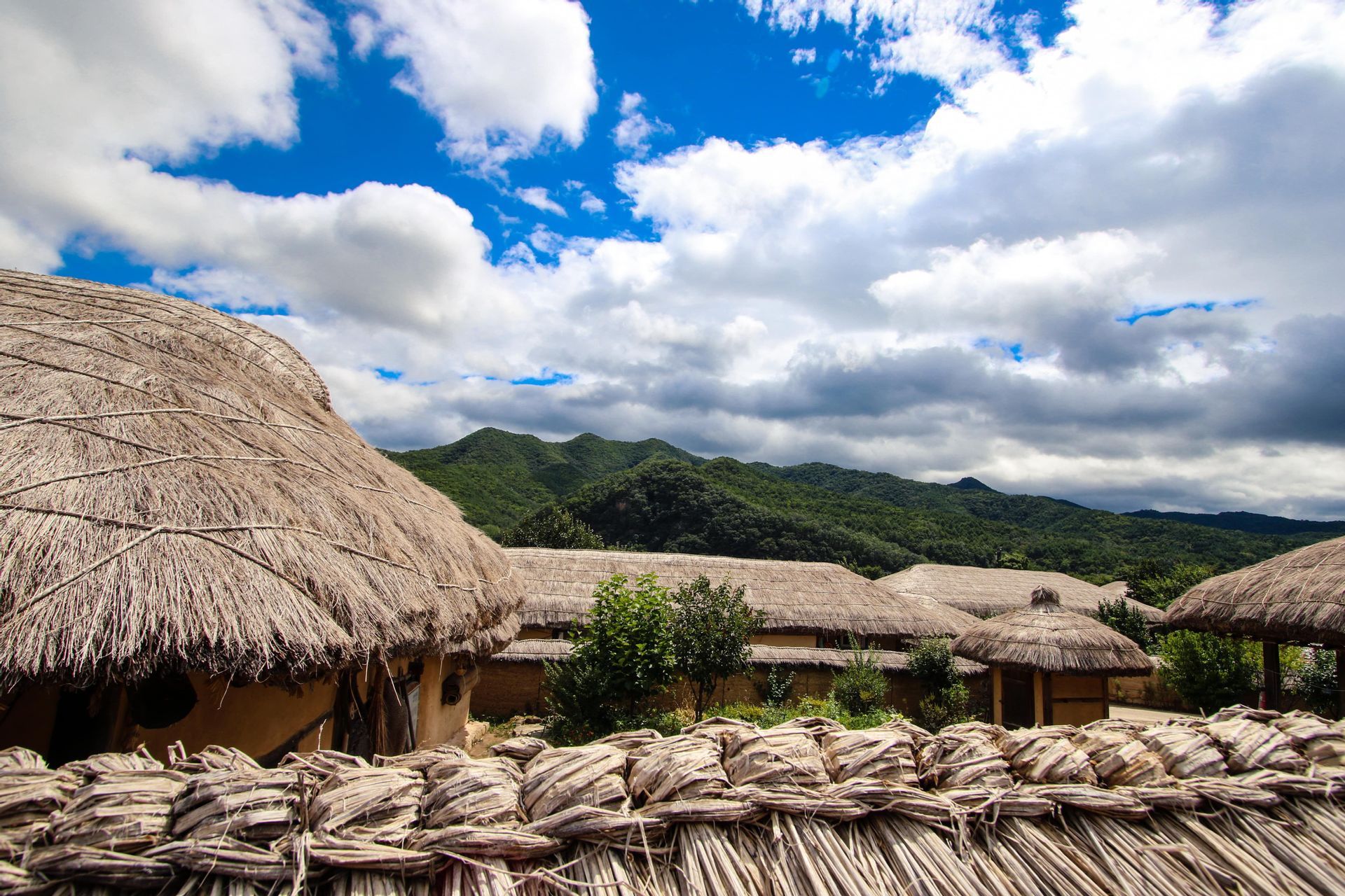 A village of traditional huts with thatched roofs sits at the base of green mountains under a partly cloudy sky.
