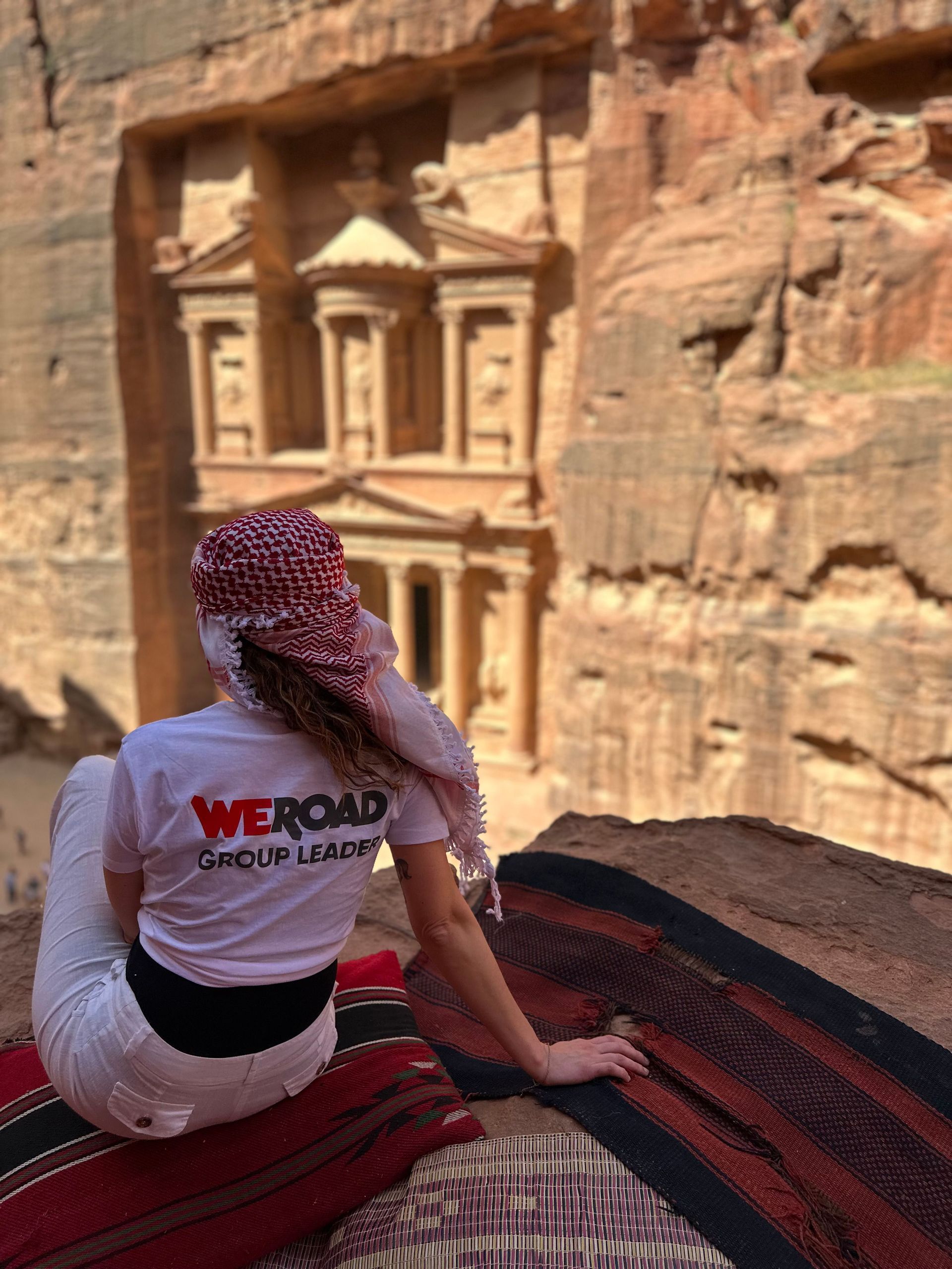 Un capogruppo WeRoad con una kefiah siede su una sporgenza che si affaccia su un antico edificio scavato in una parete di roccia rossa.