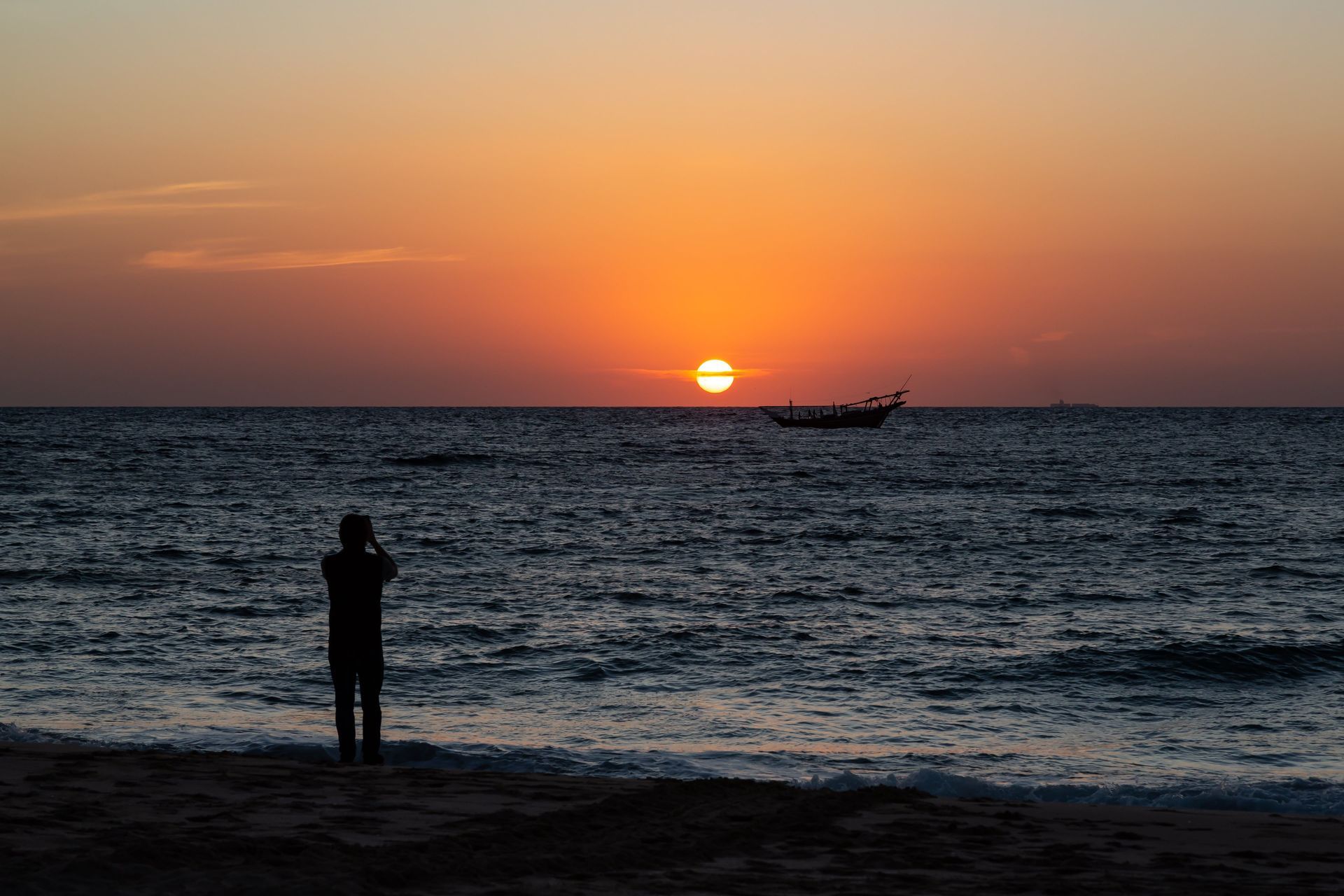 Una persona in silhouette su una spiaggia fotografa il tramonto arancione sull'oceano, con una barca all'orizzonte.