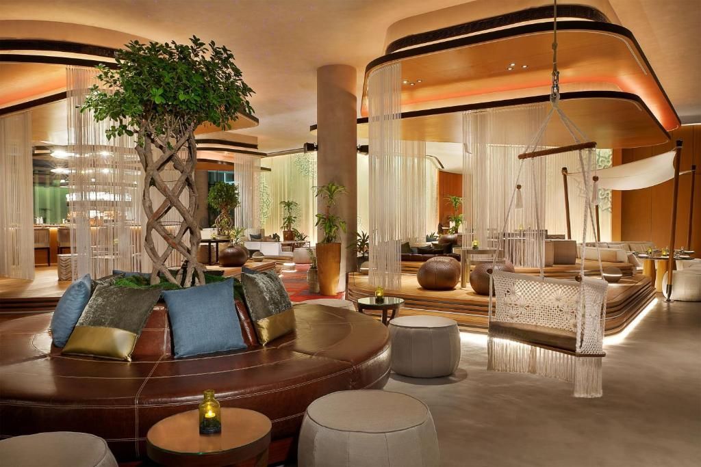 L'interno di una lounge moderna con un divano circolare in pelle marrone attorno a un albero dal tronco intrecciato e un'altalena sospesa.