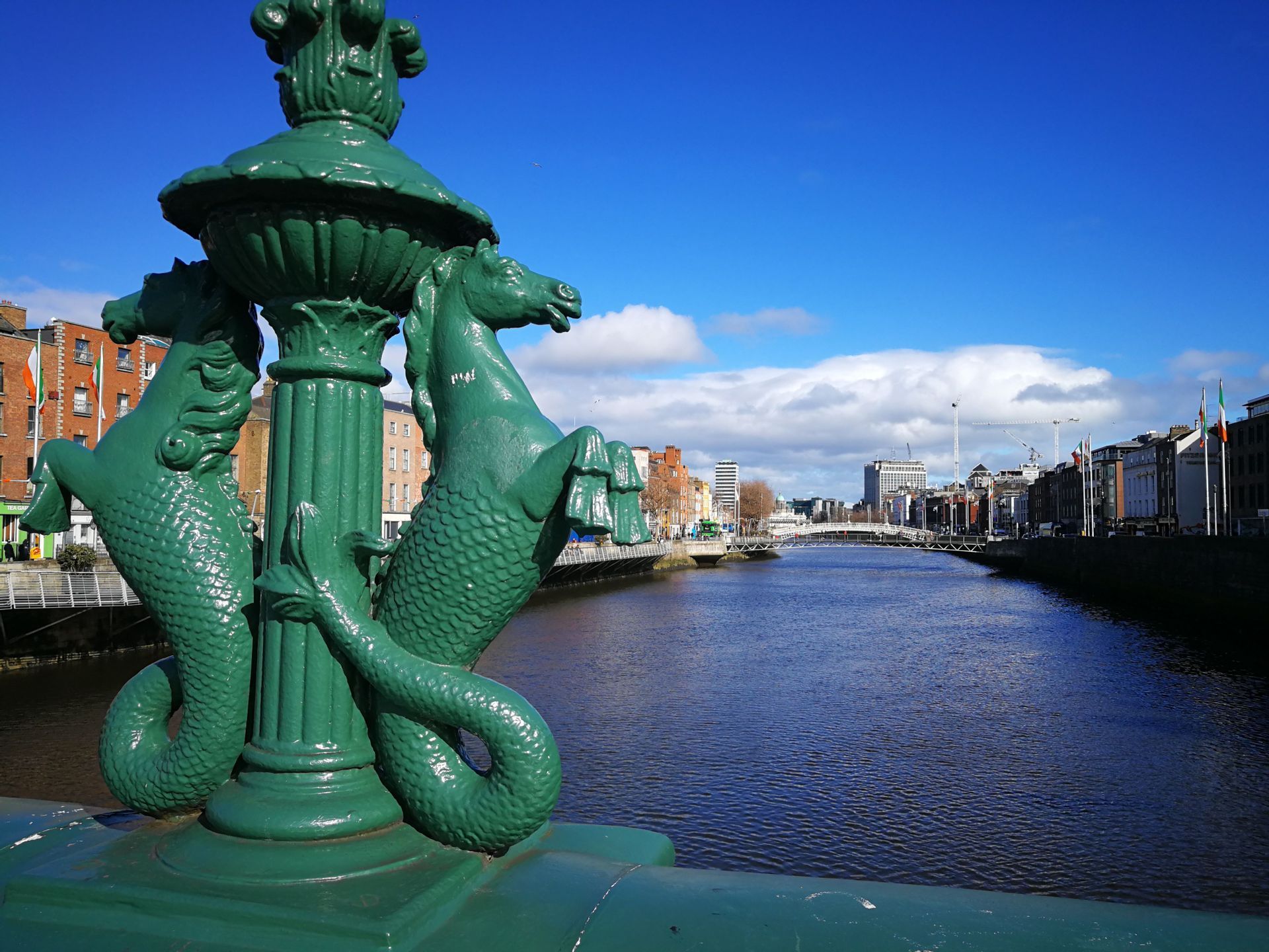 Un farol verde ornamentado con esculturas de hipocampos en un puente con vistas a un río que atraviesa una ciudad bajo un cielo azul.