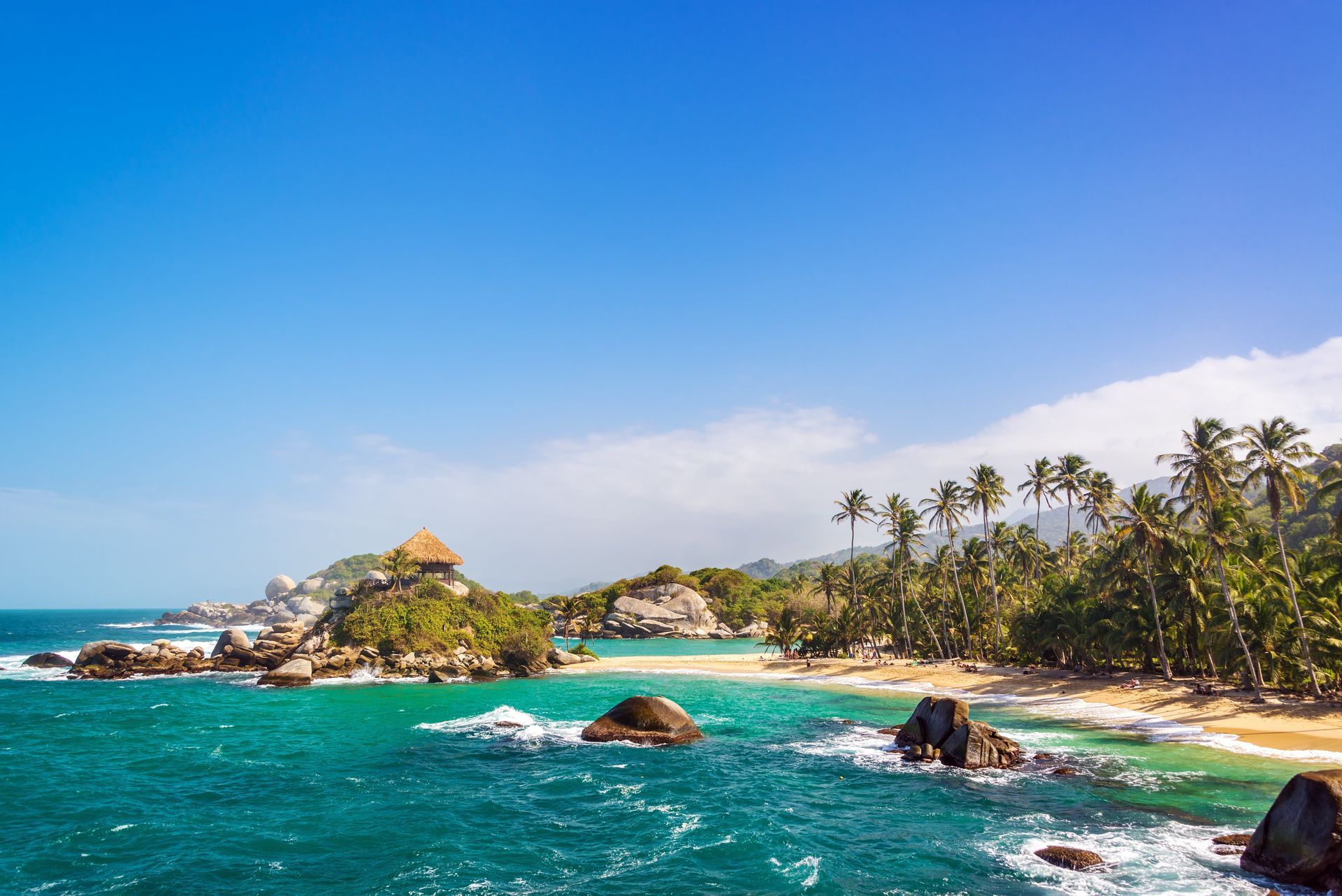 A hut with a thatched roof sits on a rocky outcrop in turquoise waters, next to a sandy beach lined with palm trees under a blue sky.