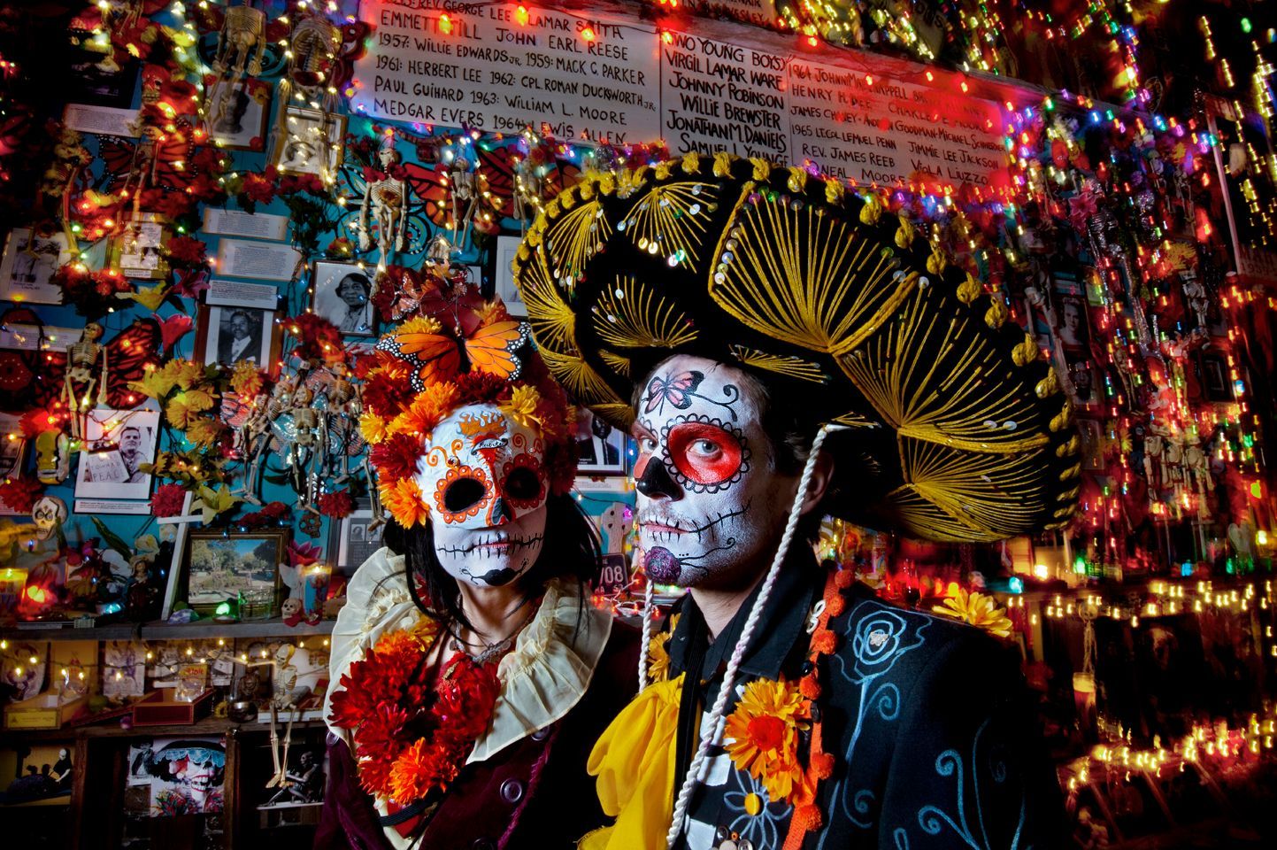 Dos personas con disfraces del Día de Muertos y maquillaje de calavera de azúcar posan frente a una pared decorada con luces y fotos.