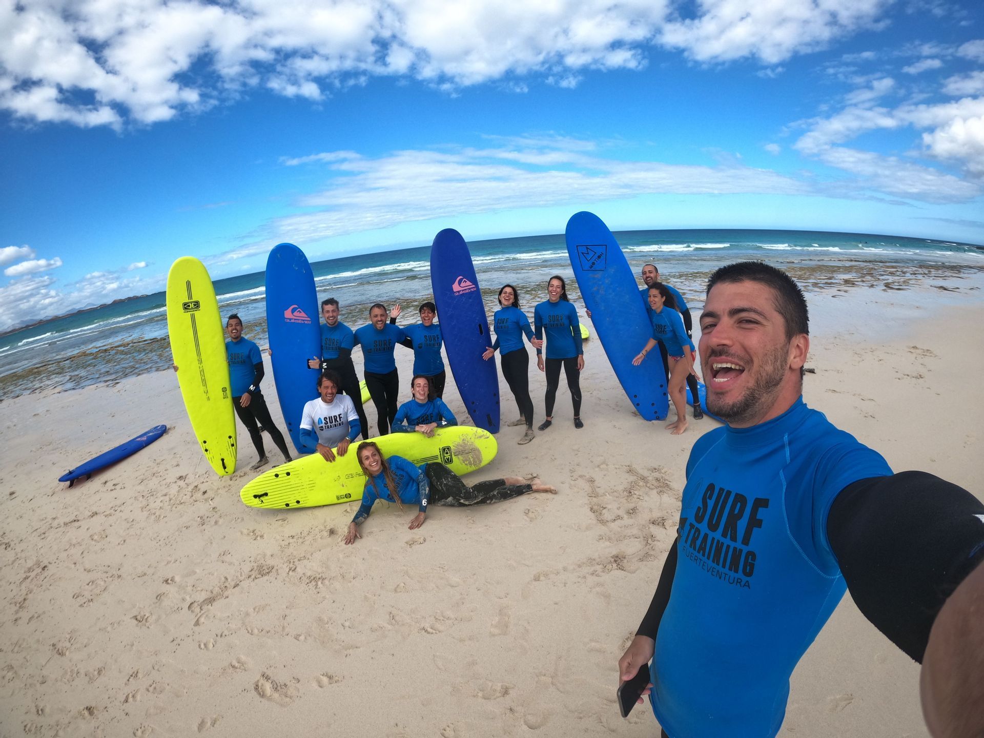 Un gruppo WeRoad si scatta un selfie su una spiaggia sabbiosa, tutti con mute blu abbinate e in posa con le loro tavole da surf.