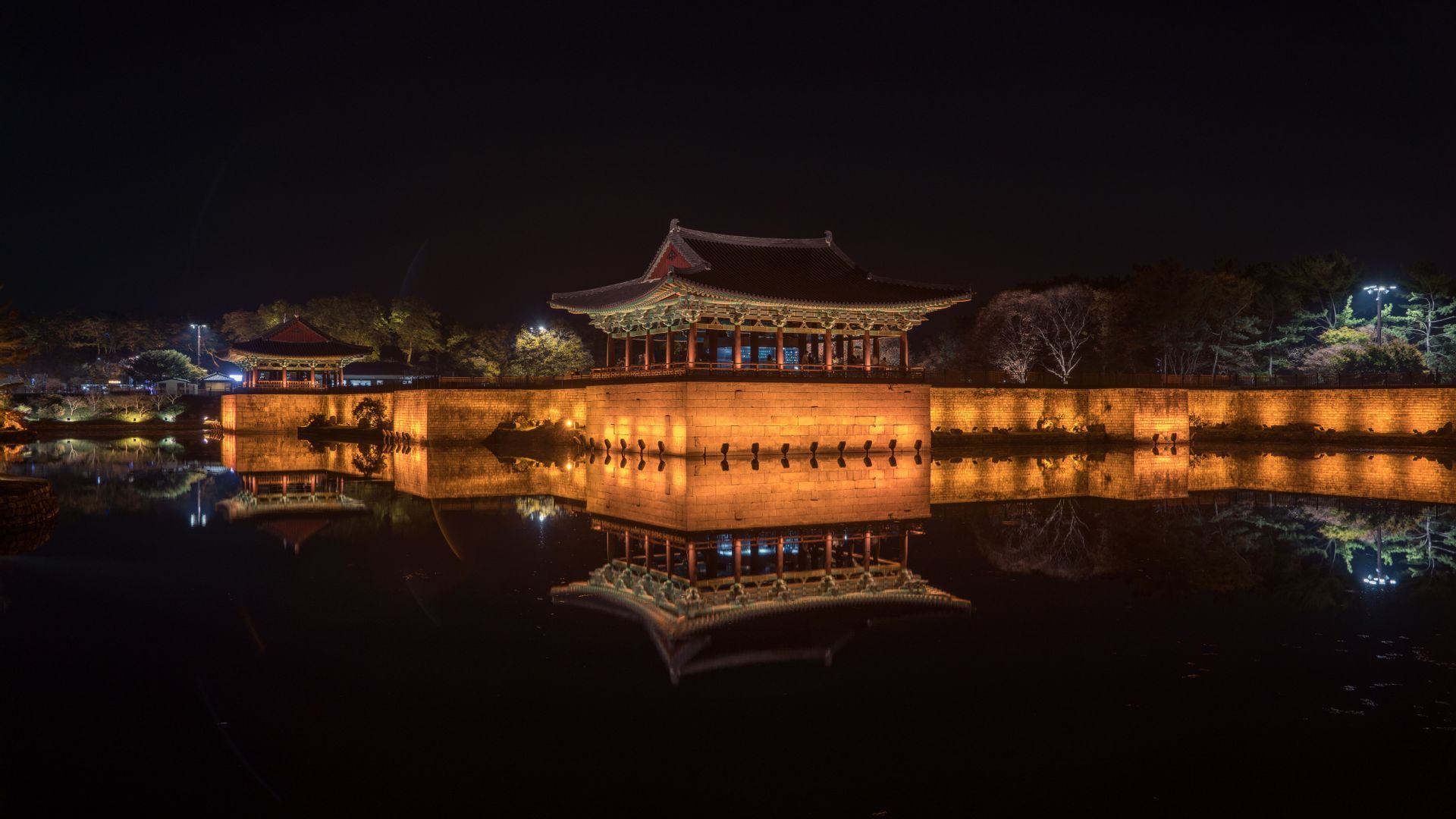 Ein traditioneller Pavillon und eine Steinmauer, nachts beleuchtet, werfen eine perfekte Spiegelung auf das ruhige Wasser darunter.