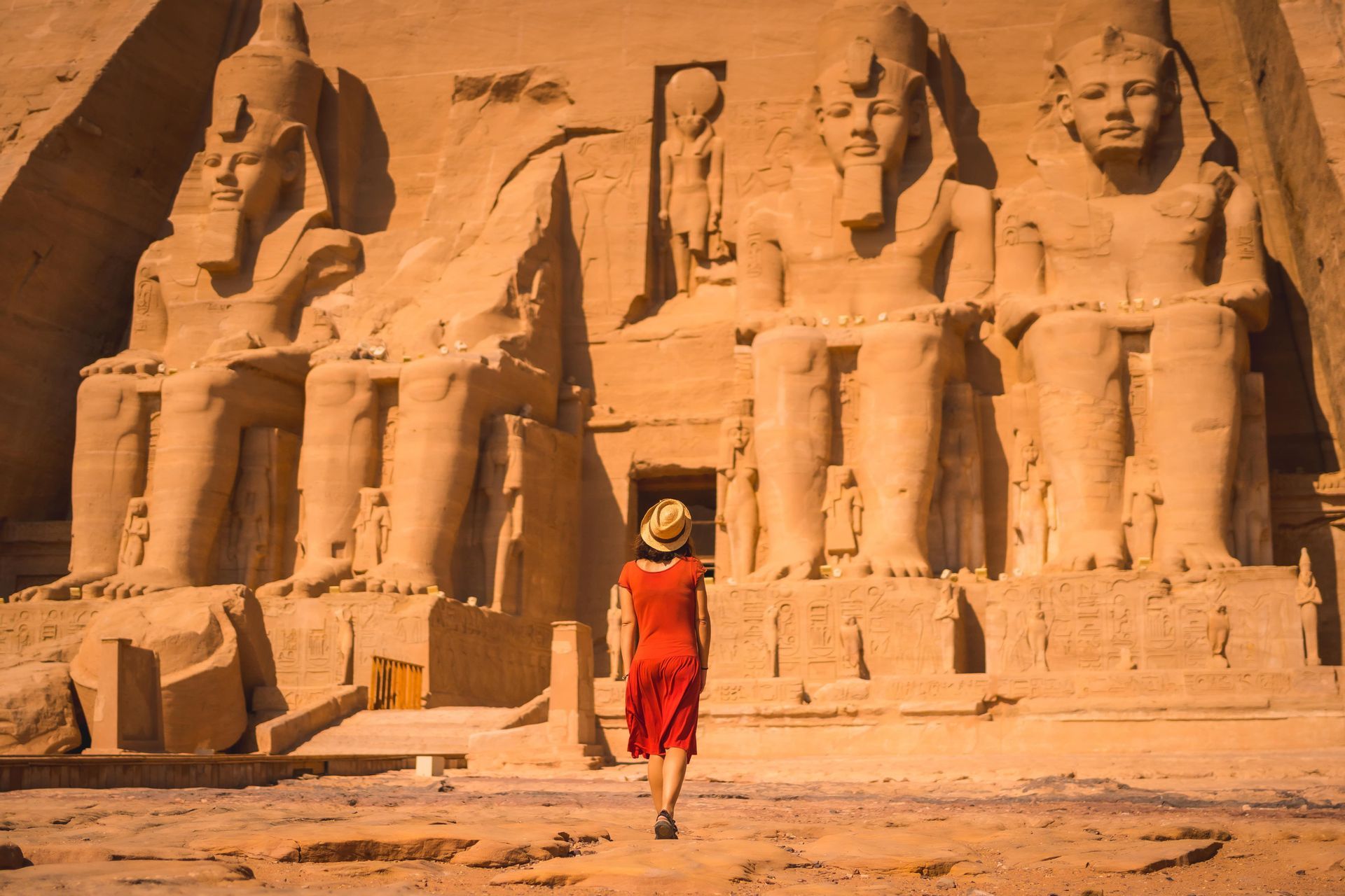 Una donna in abito rosso e cappello di paglia cammina verso un grande tempio antico scolpito in una scogliera con colossali statue sedute.