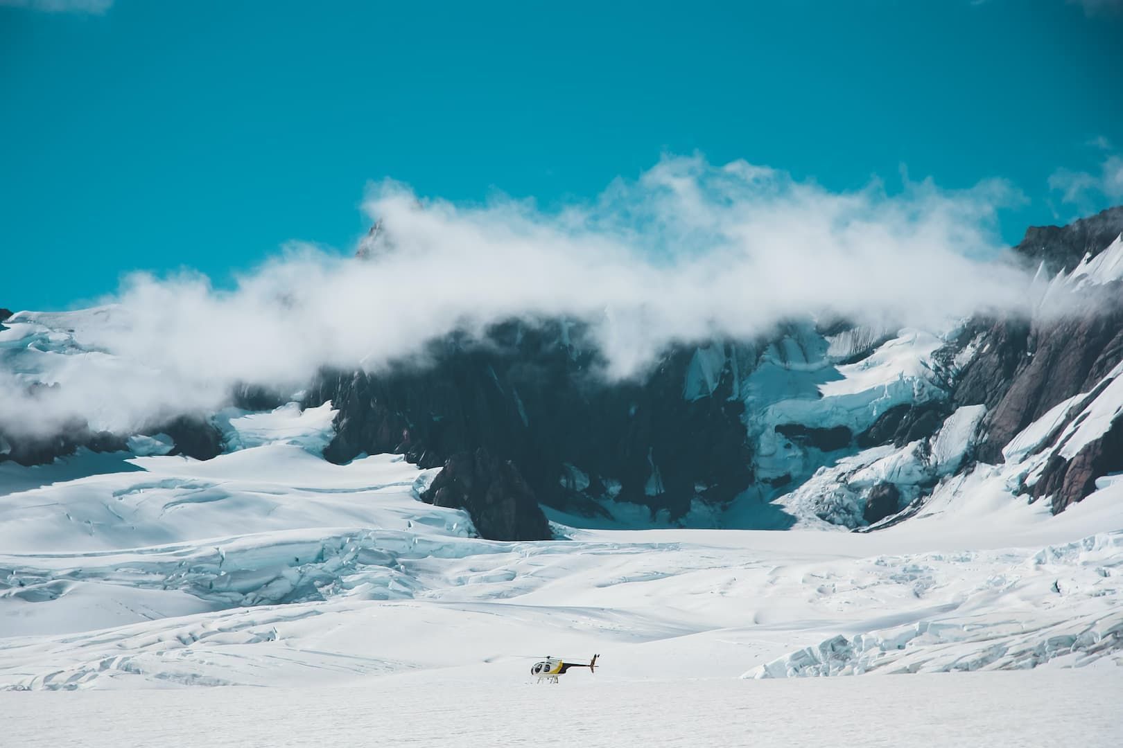Ein Hubschrauber ruht auf einem riesigen Gletscher am Fuße schneebedeckter Berge unter blauem Himmel.