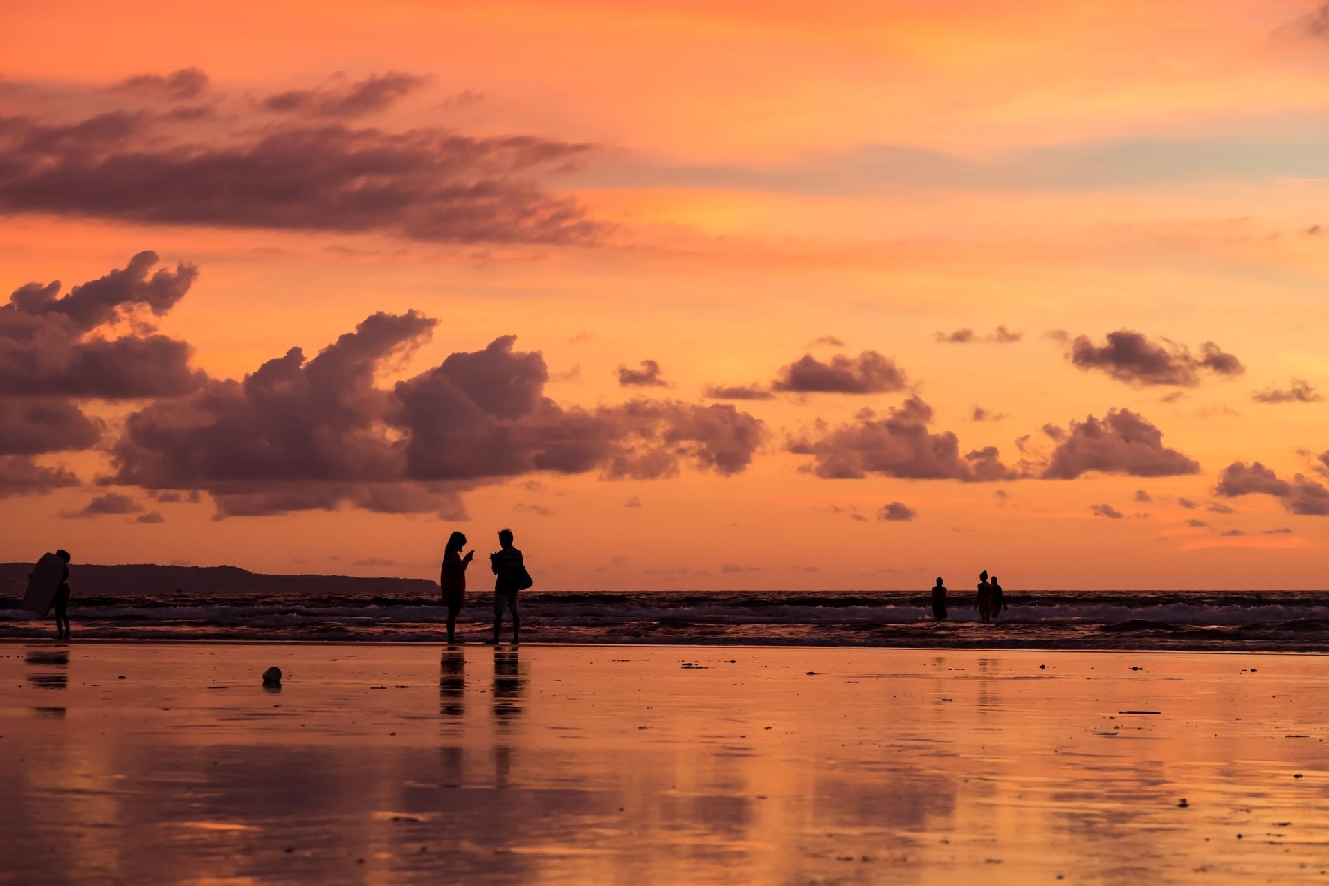 Silhouetten einer WeRoad Gruppenreise an einem nassen Strand bei Sonnenuntergang, mit einem orangefarbenen Himmel und Wolken, die sich im Sand spiegeln.