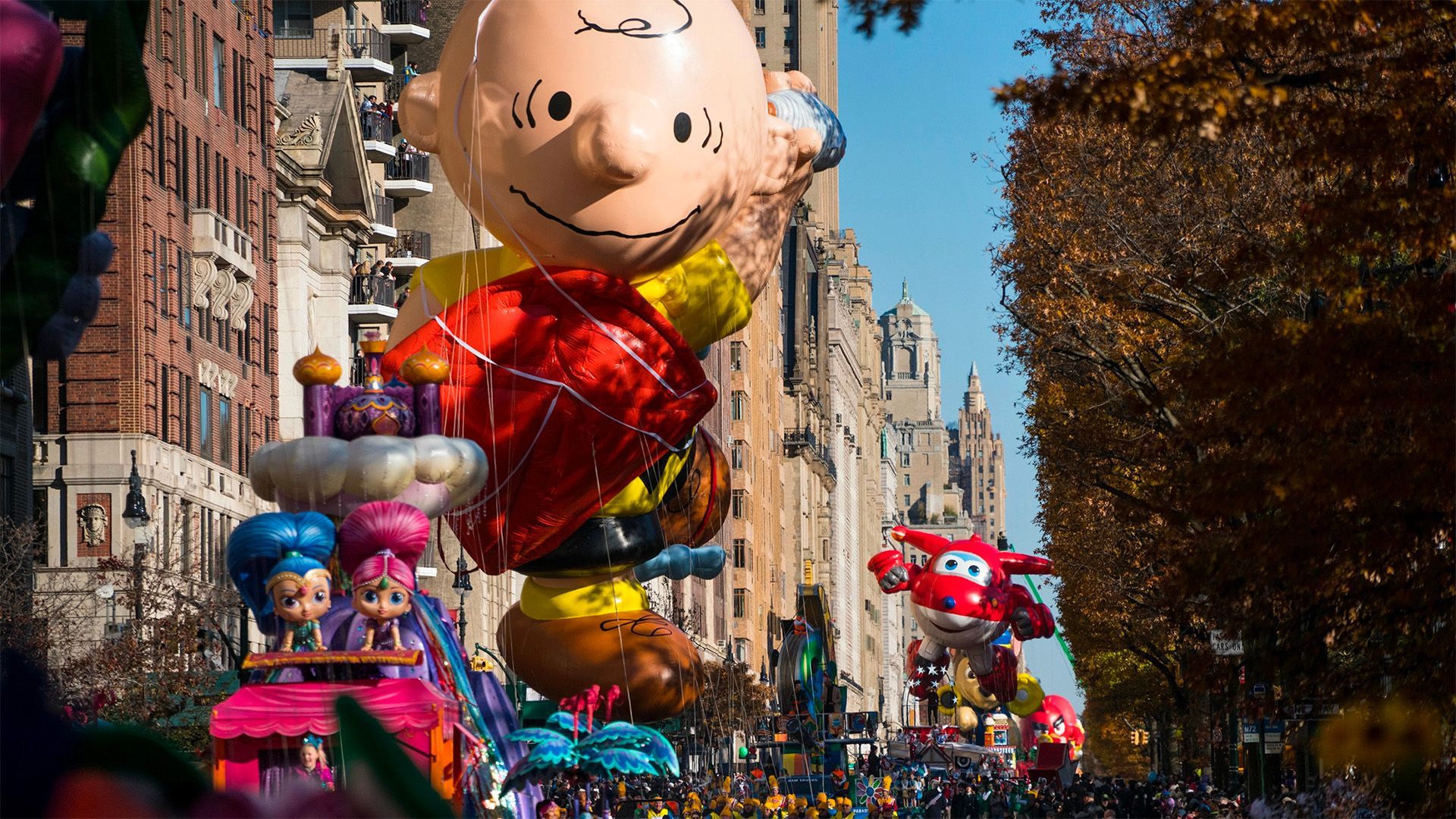 Un gigantesco pallone di Charlie Brown e altri carri a tema avanzano lungo una via cittadina durante una parata sotto un cielo azzurro e limpido.