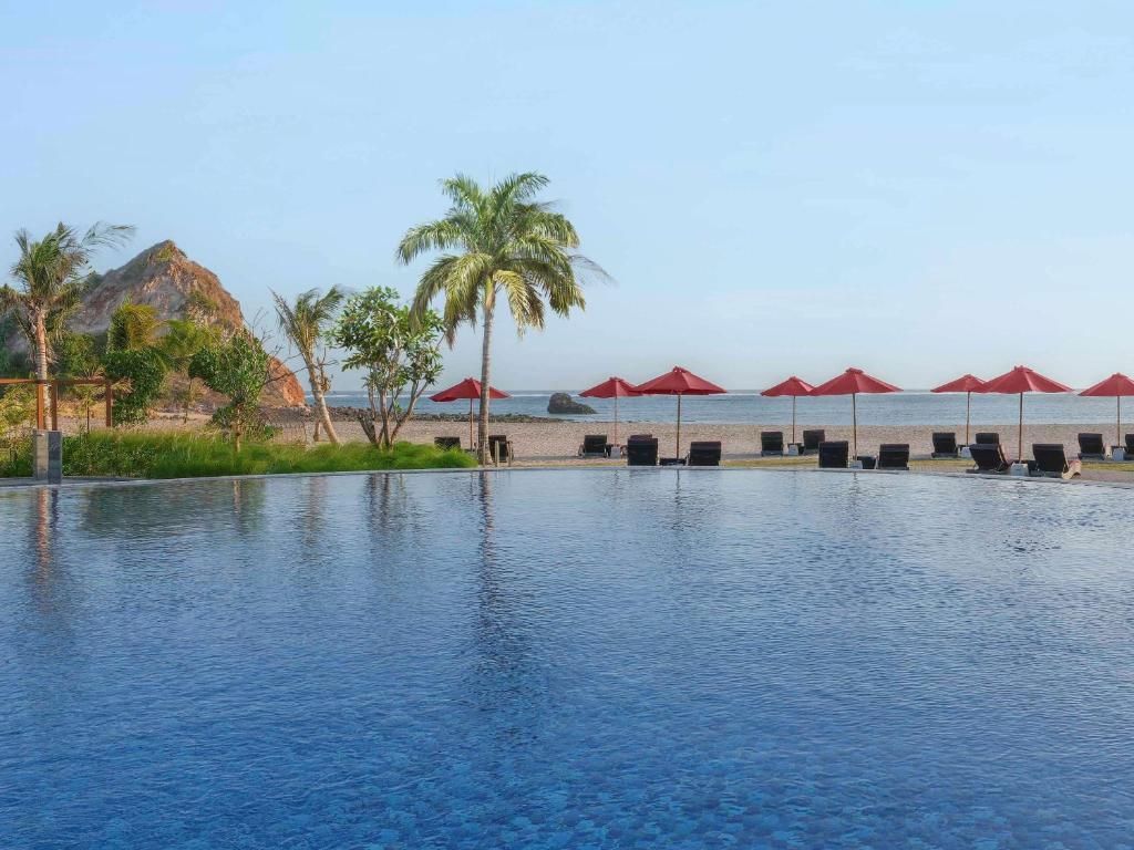 Ein großer Infinity-Pool überblickt einen Sandstrand mit roten Sonnenschirmen, Liegestühlen und Palmen unter klarem Himmel.