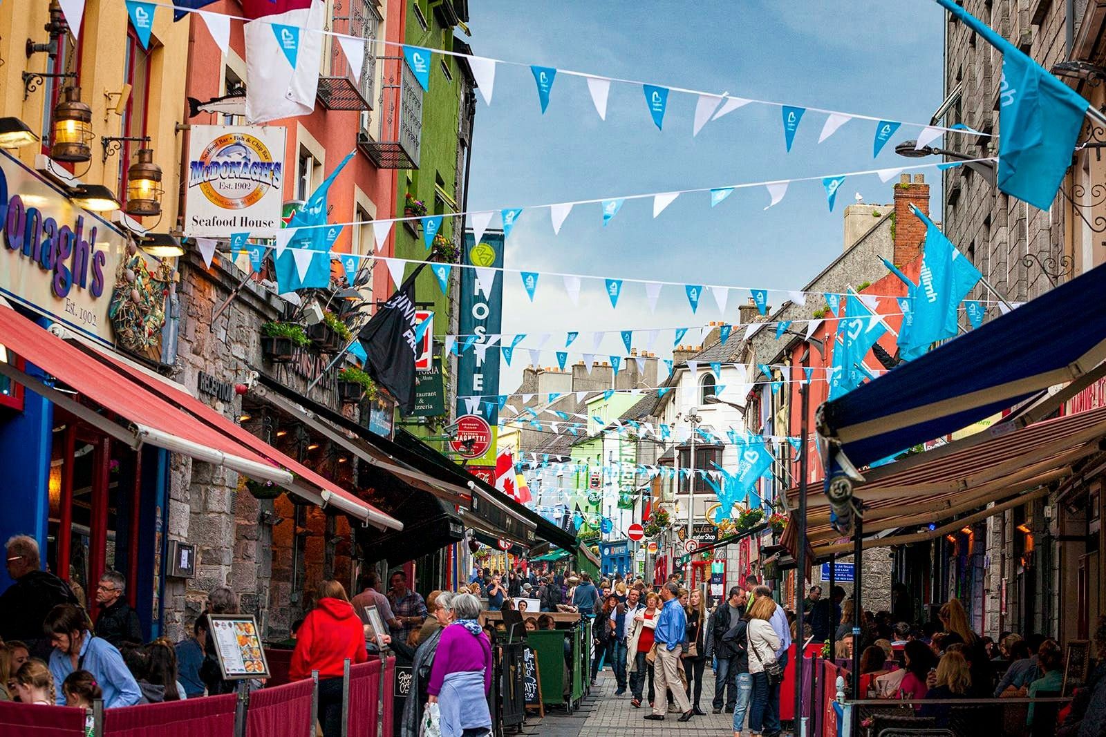 Una calle peatonal concurrida, flanqueada por edificios y restaurantes coloridos, decorada con guirnaldas de banderas azules y blancas.