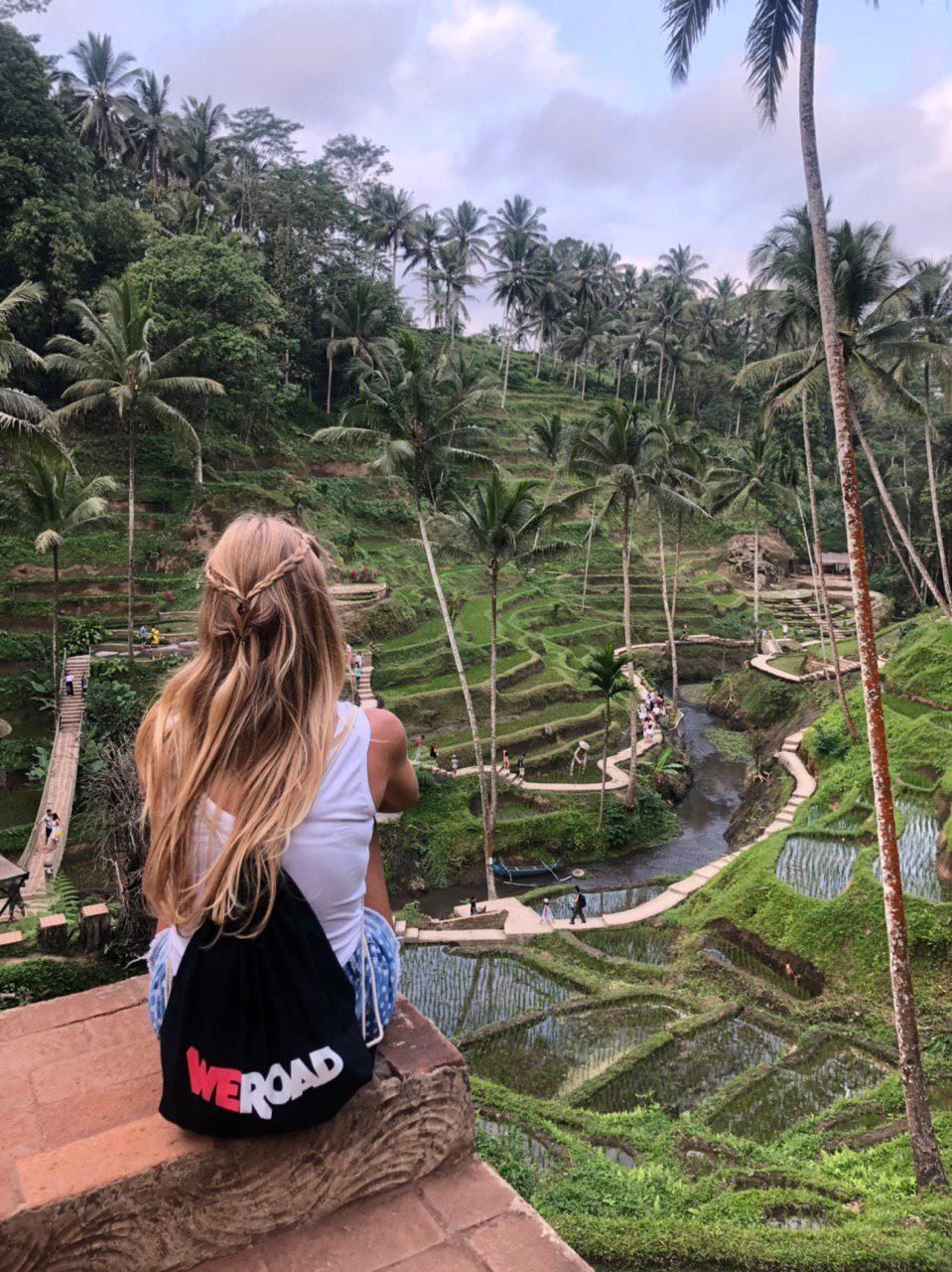 Una donna con una borsa WeRoad siede su un cornicione che si affaccia su una valle con risaie terrazzate verdi e alte palme.