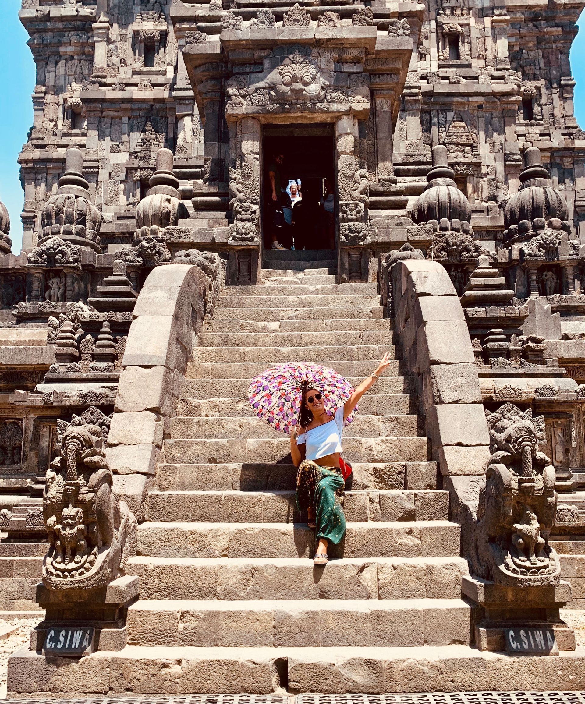 A woman holding a colorful patterned umbrella sits on the wide stone steps of an ornate ancient temple.